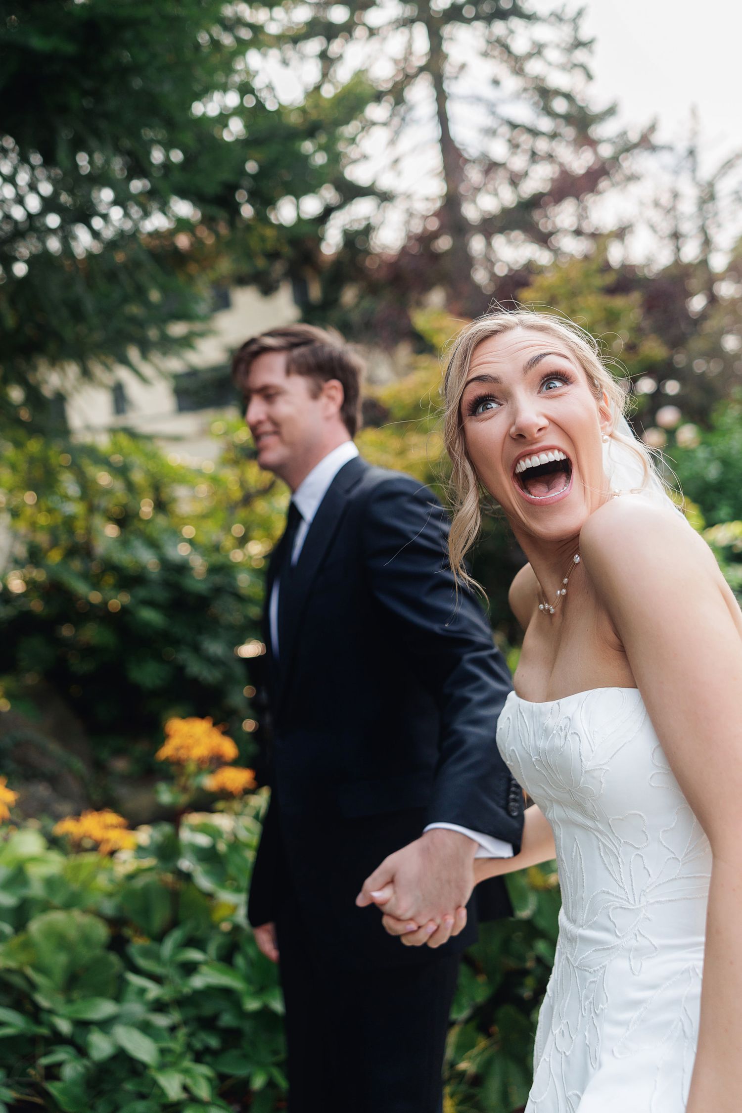 Joyful bride in white strapless gown laughing outdoors, holding hands with groom in black suit amid lush garden greenery.