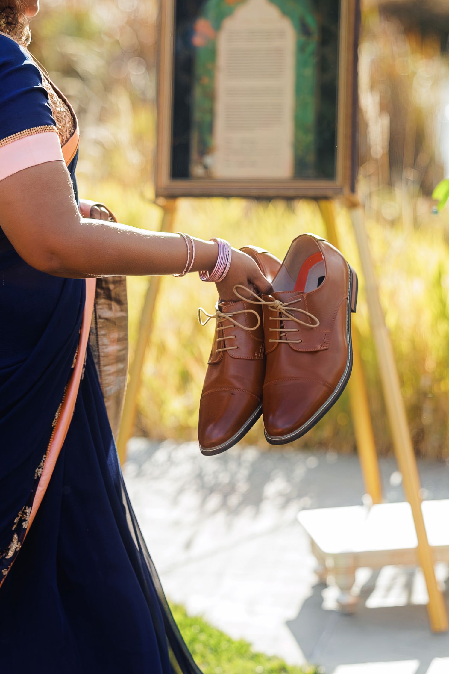 Woman in blue saree holding brown leather lace-up oxford shoes at an outdoor Indian wedding ceremony.
