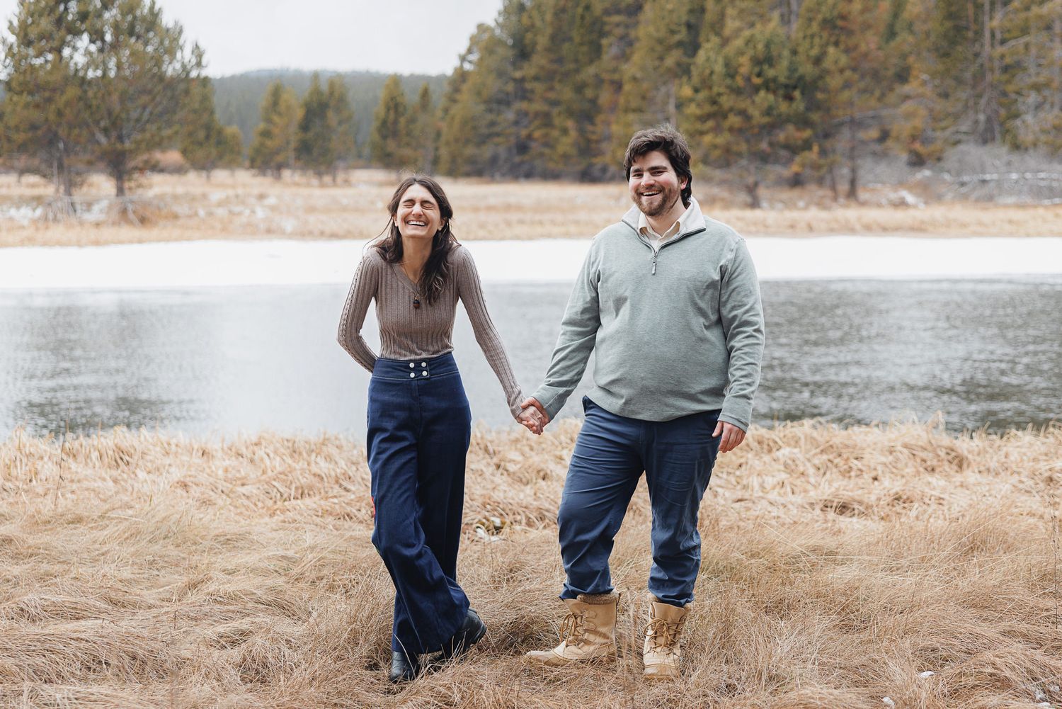 A couple walks together through golden dried grass near a serene lake with evergreen trees in the background.
