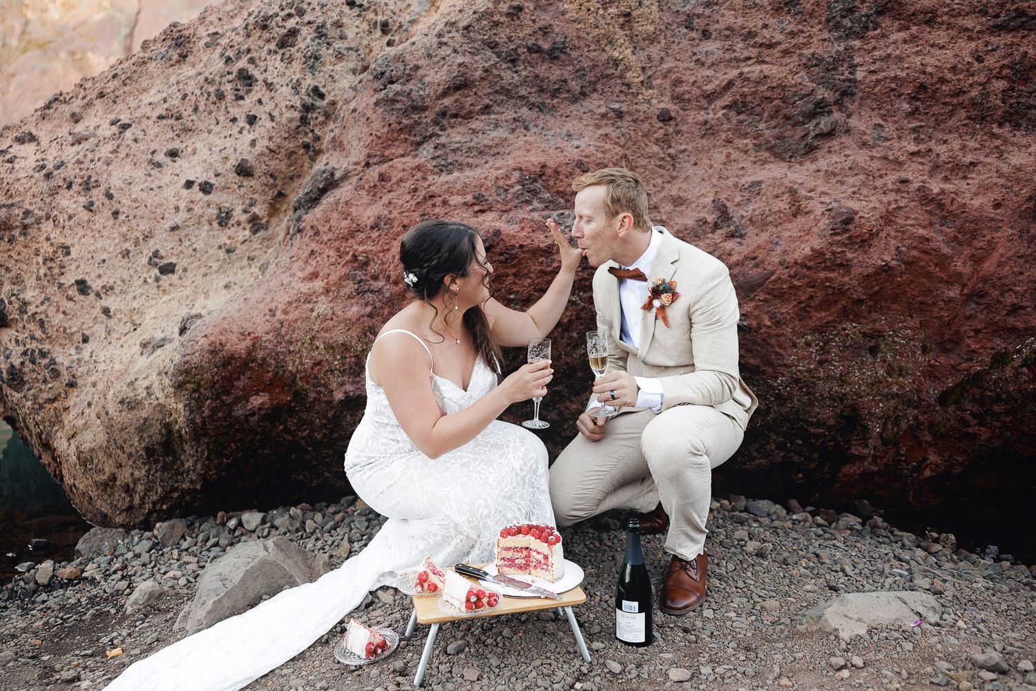 Bride in a white gown and groom in a beige suit toast with champagne beside a small cake and wine bottle against red volcanic rock.