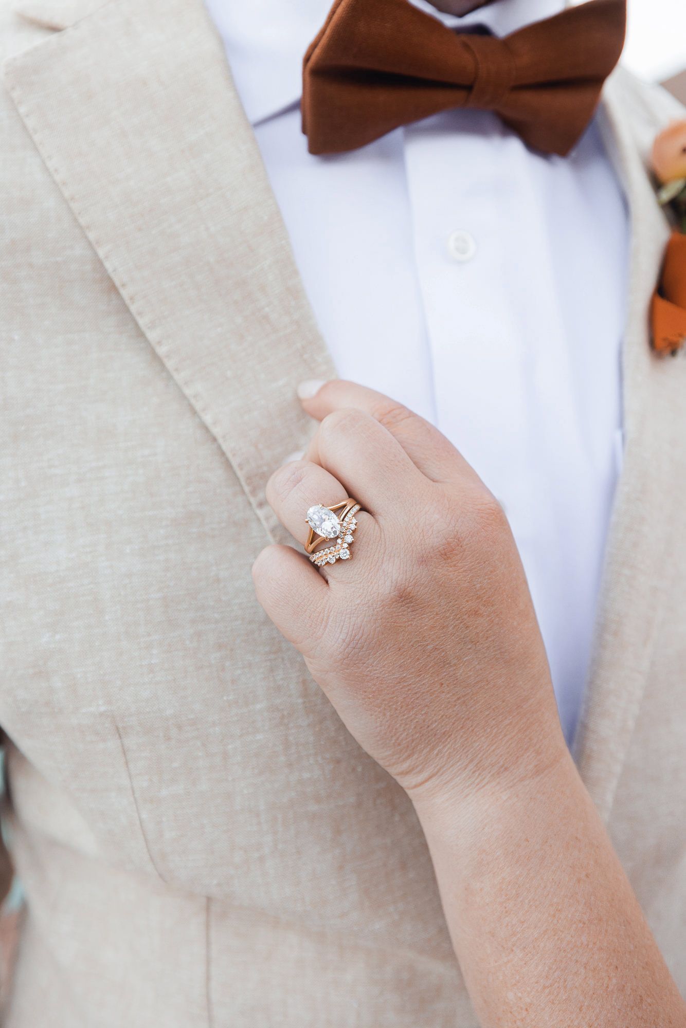 Close-up of a hand with a diamond engagement ring and wedding band resting on a beige linen suit jacket with a rust bow tie.