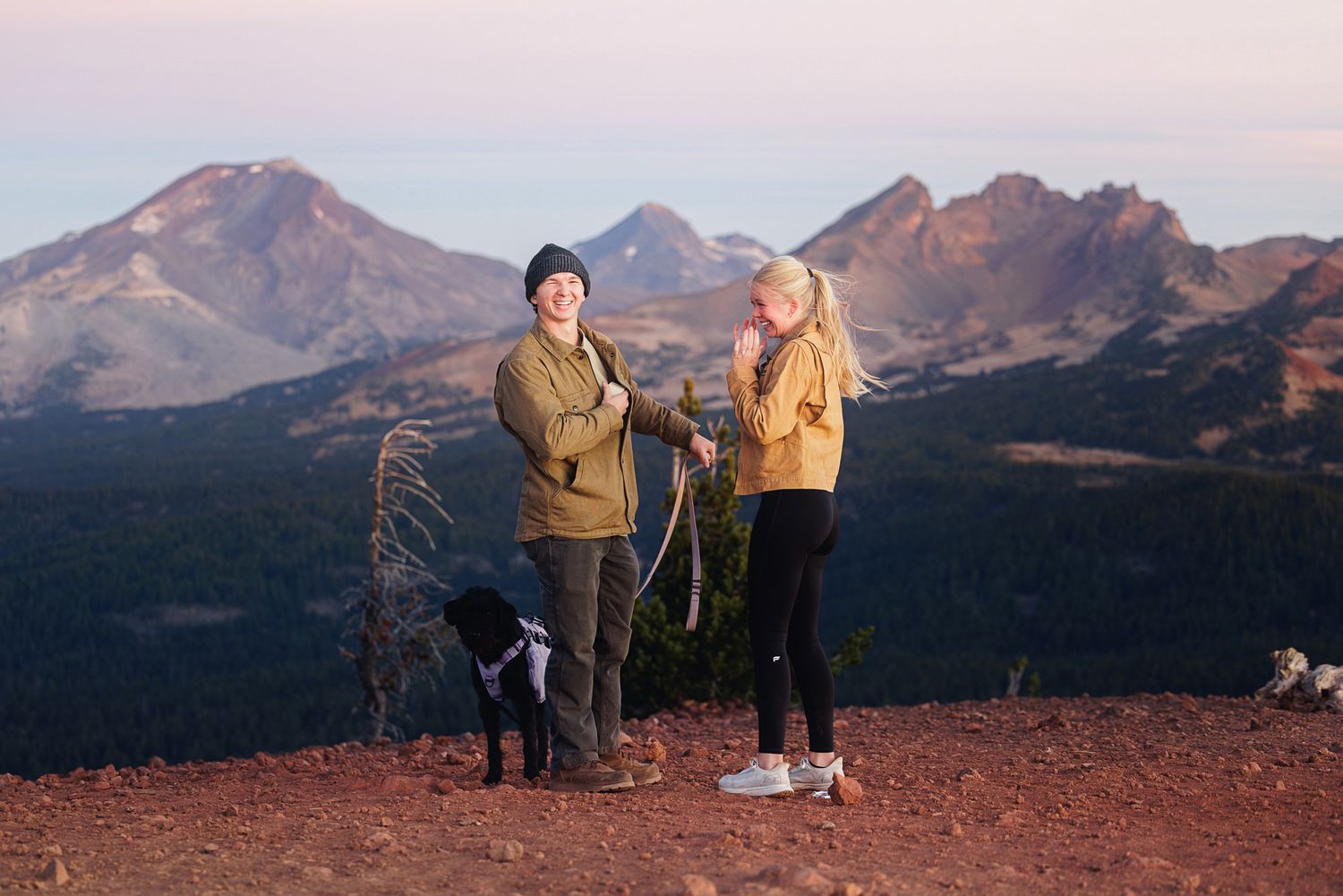 Two hikers in warm jackets stand with a black dog on a red rocky summit at sunset, with rugged mountain peaks behind them.