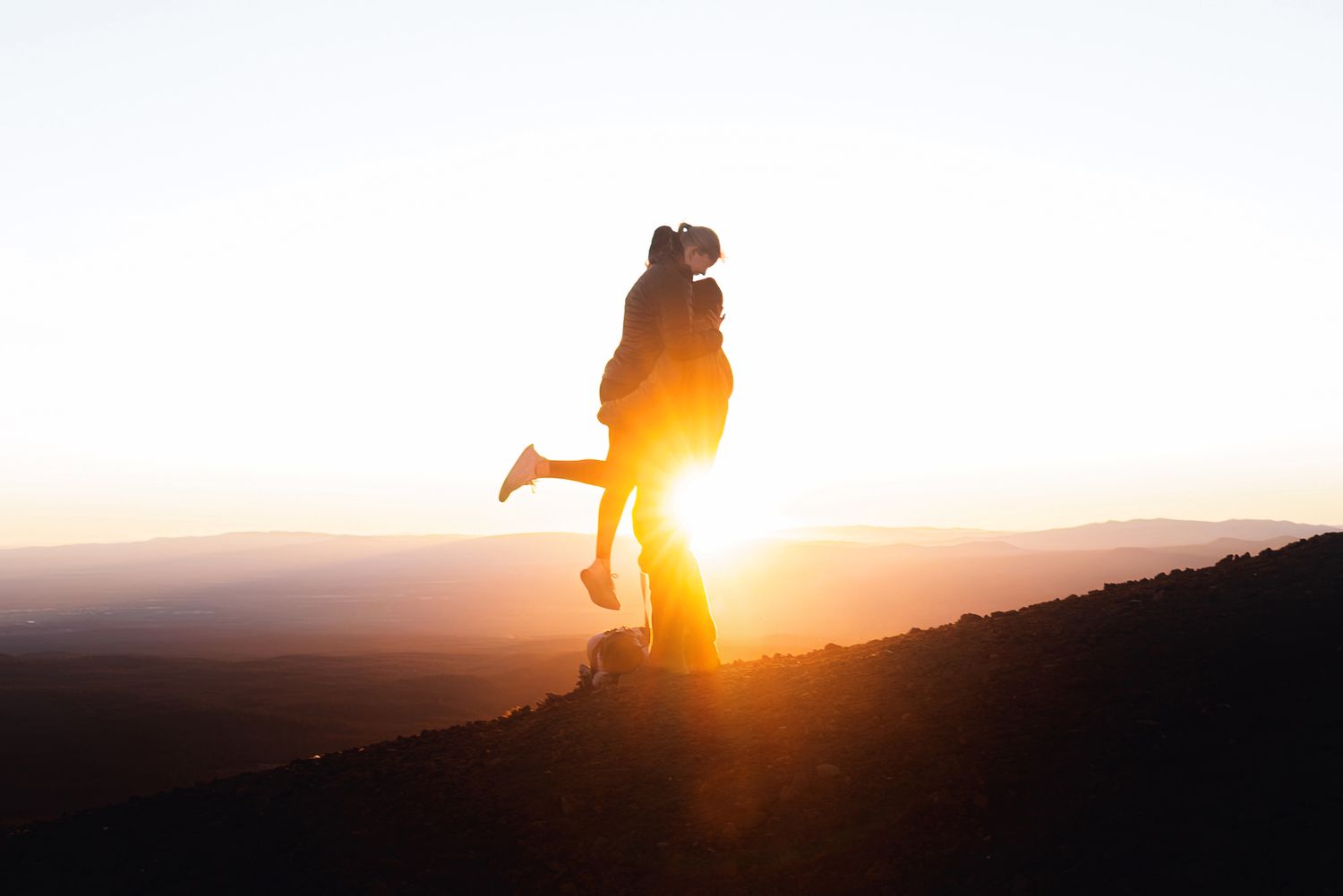 Silhouetted couple embracing on a hilltop at sunset, with one person lifted off the ground against a glowing golden sky.