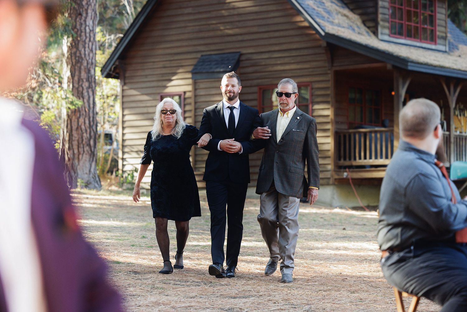Groom walks with parents toward outdoor wedding ceremony near rustic log cabin surrounded by tall trees.