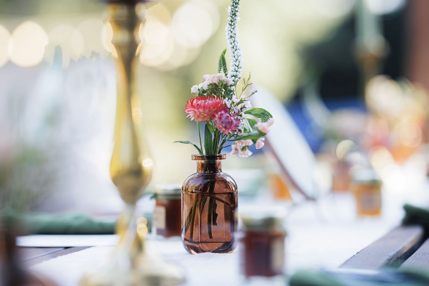 Small amber glass vase with colorful wildflowers on a blurred elegant table setting.