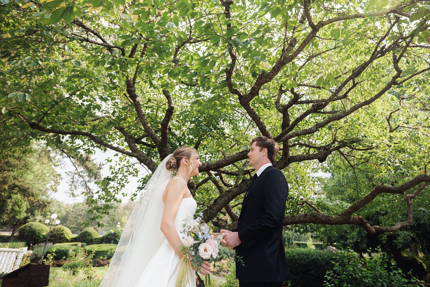 Bride and groom share a romantic moment under a lush green tree in an outdoor garden wedding setting.