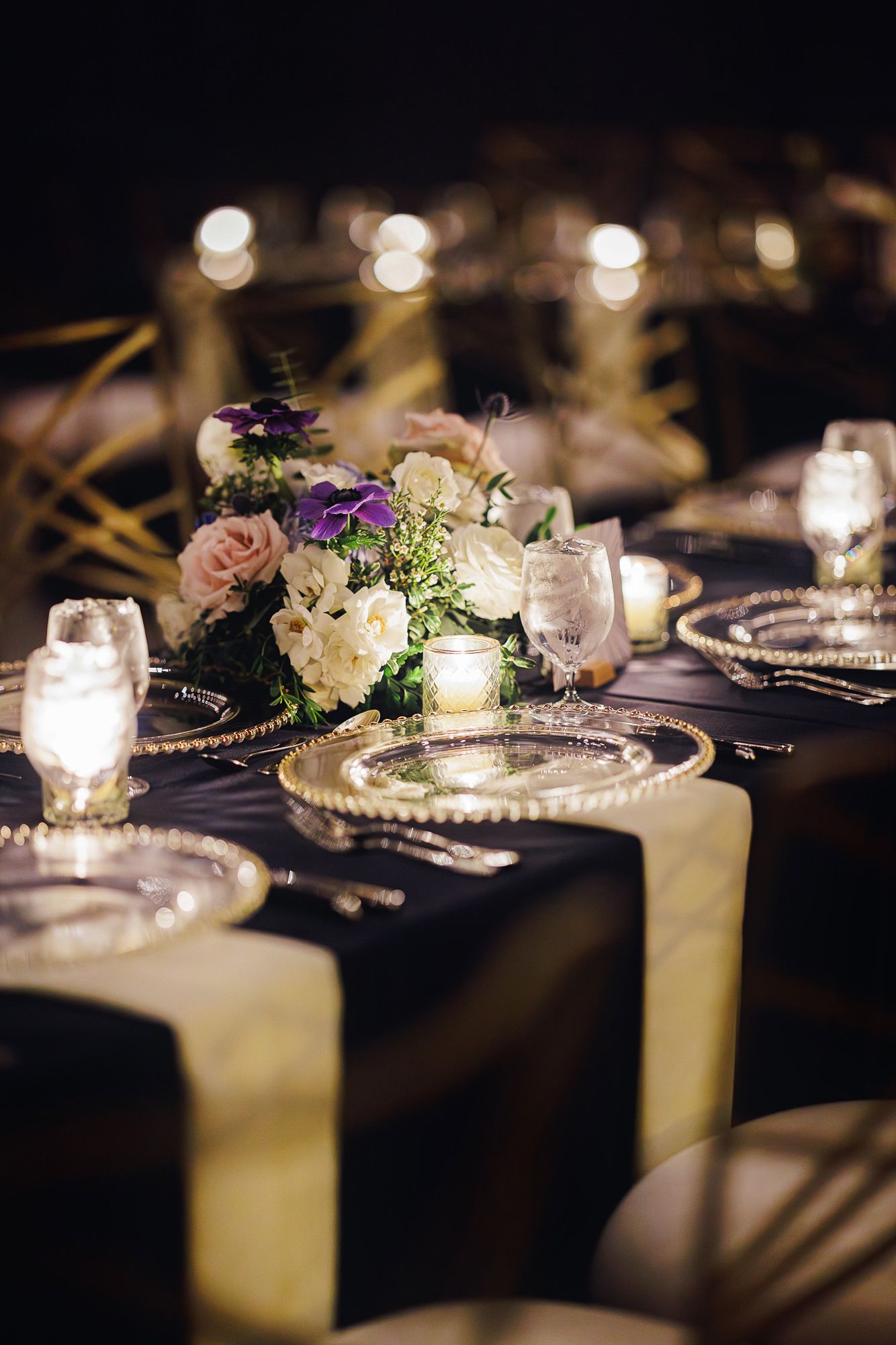Elegant wedding table setting with candlelight and floral centerpieces on dark tablecloths in moody lighting.