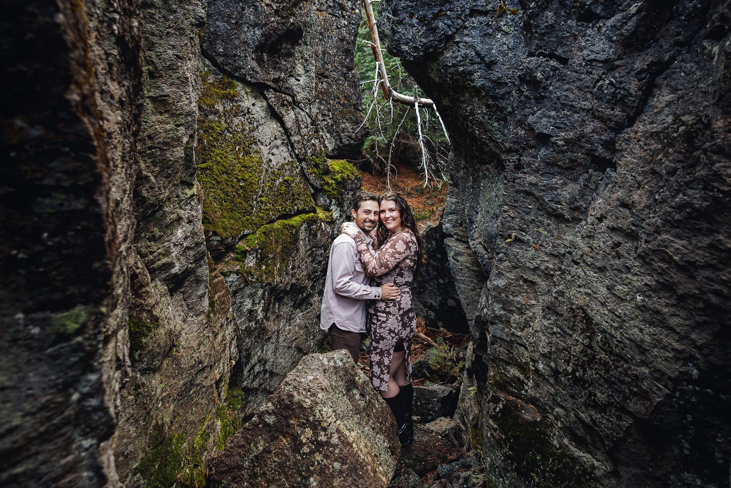 A couple embraces while standing in front of dark rocky cliffs with moss and trees in the background.