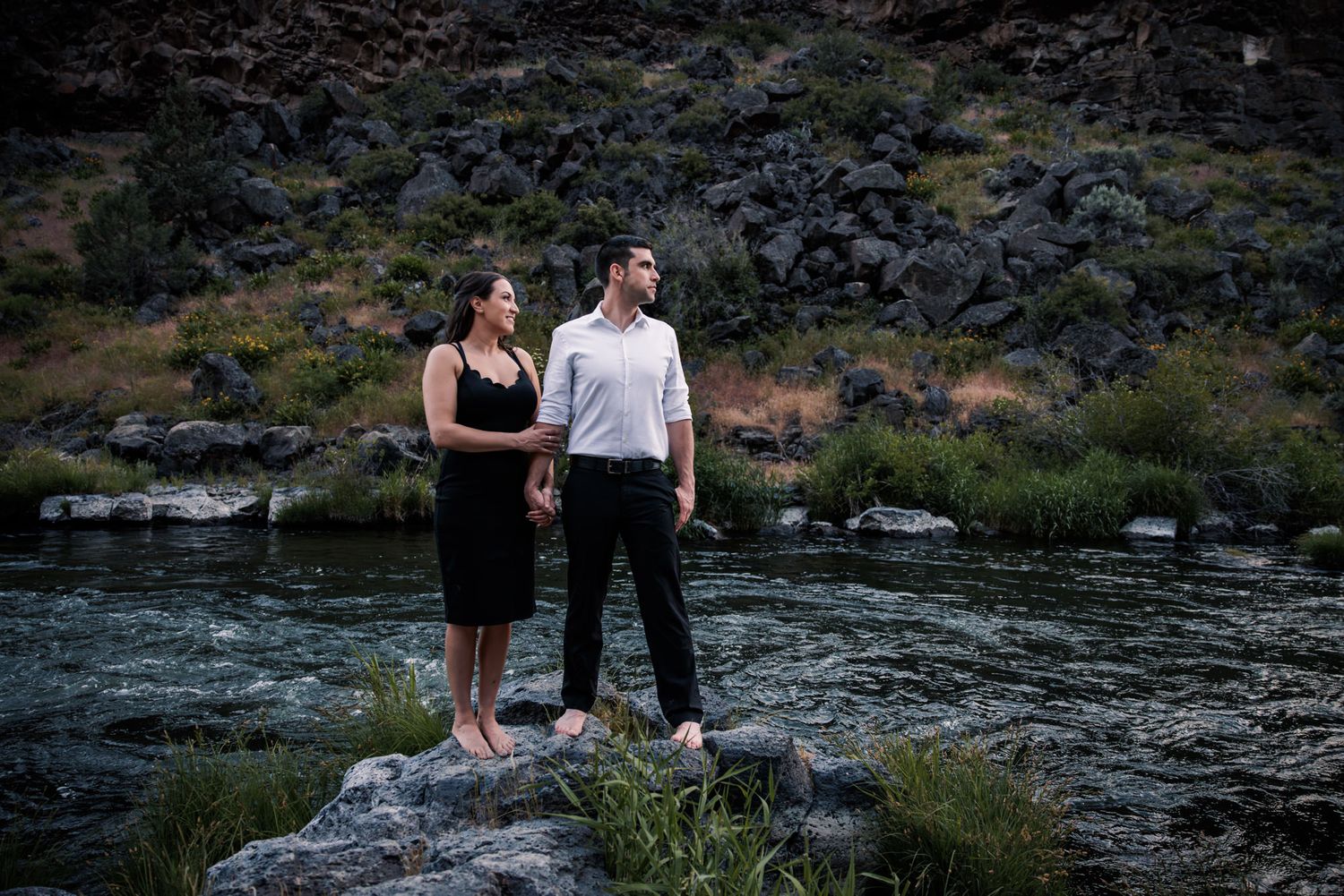 A couple stands together on rocks beside a rushing river at dusk in a scenic mountain landscape.