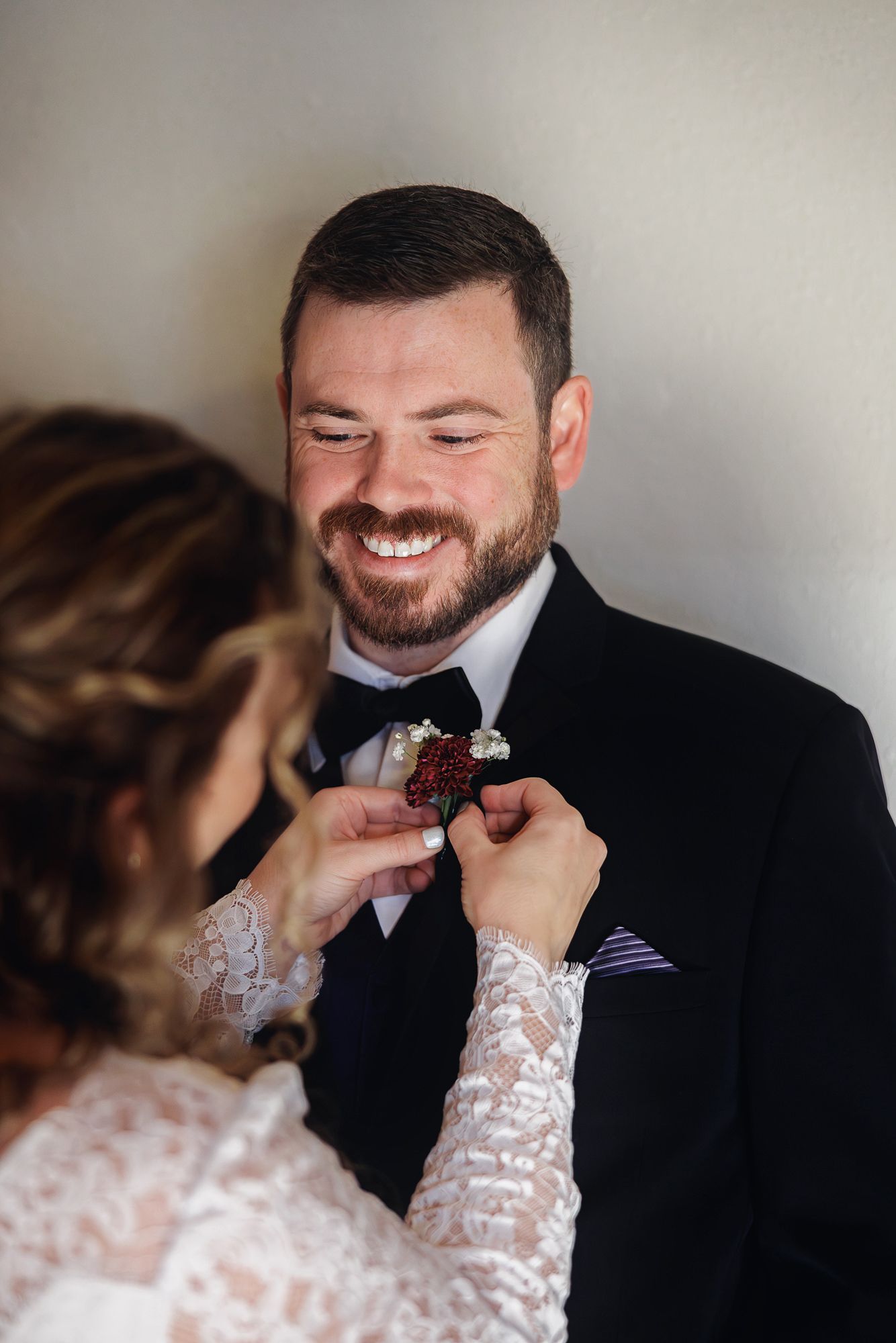 Someone wearing a black tuxedo smiles warmly while having their bow tie adjusted by hands in lace sleeves.