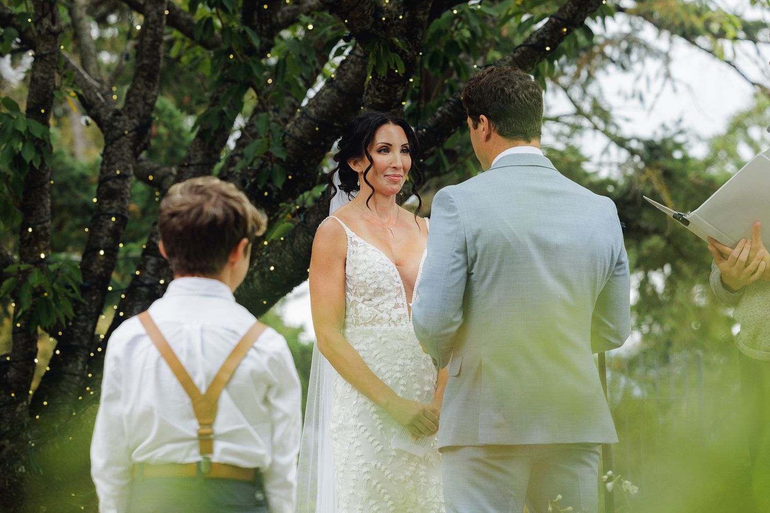 A couple in wedding attire walks together on a garden path with lush greenery and trees in the background.