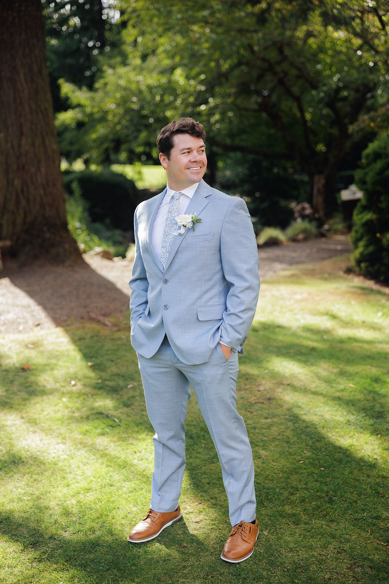 A person in a light blue suit and brown shoes poses in a sunny garden setting with trees in the background.