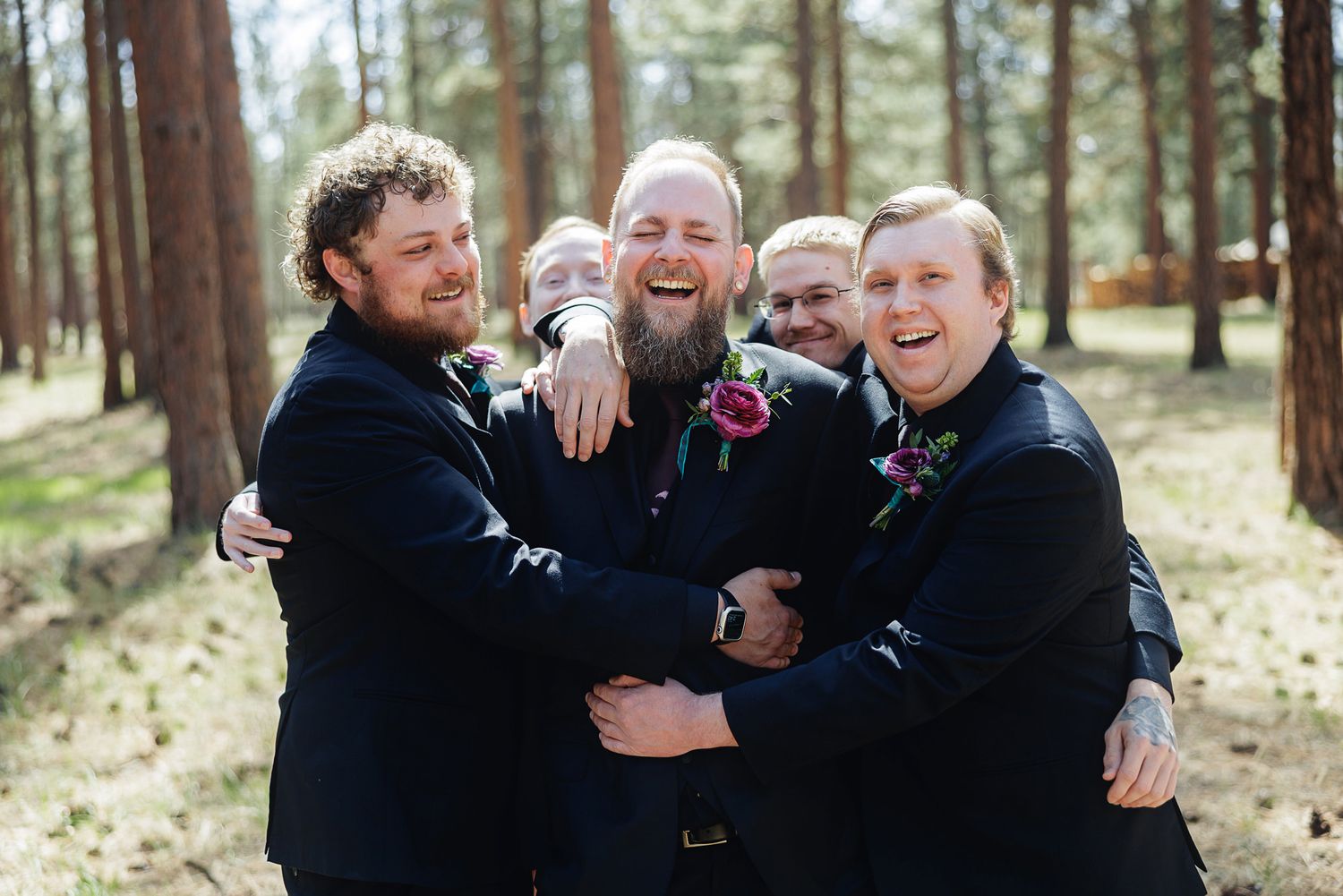 Groomsmen in navy suits hugging and laughing together in a pine forest during a wedding celebration.