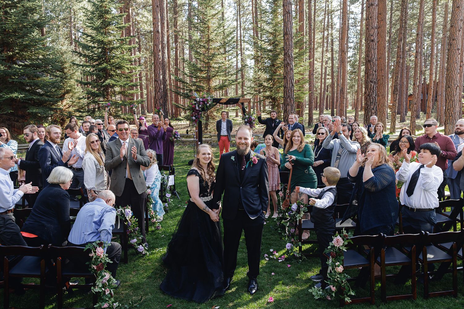 Couple walks down forest aisle in black wedding attire, guests applaud at outdoor woodland ceremony.