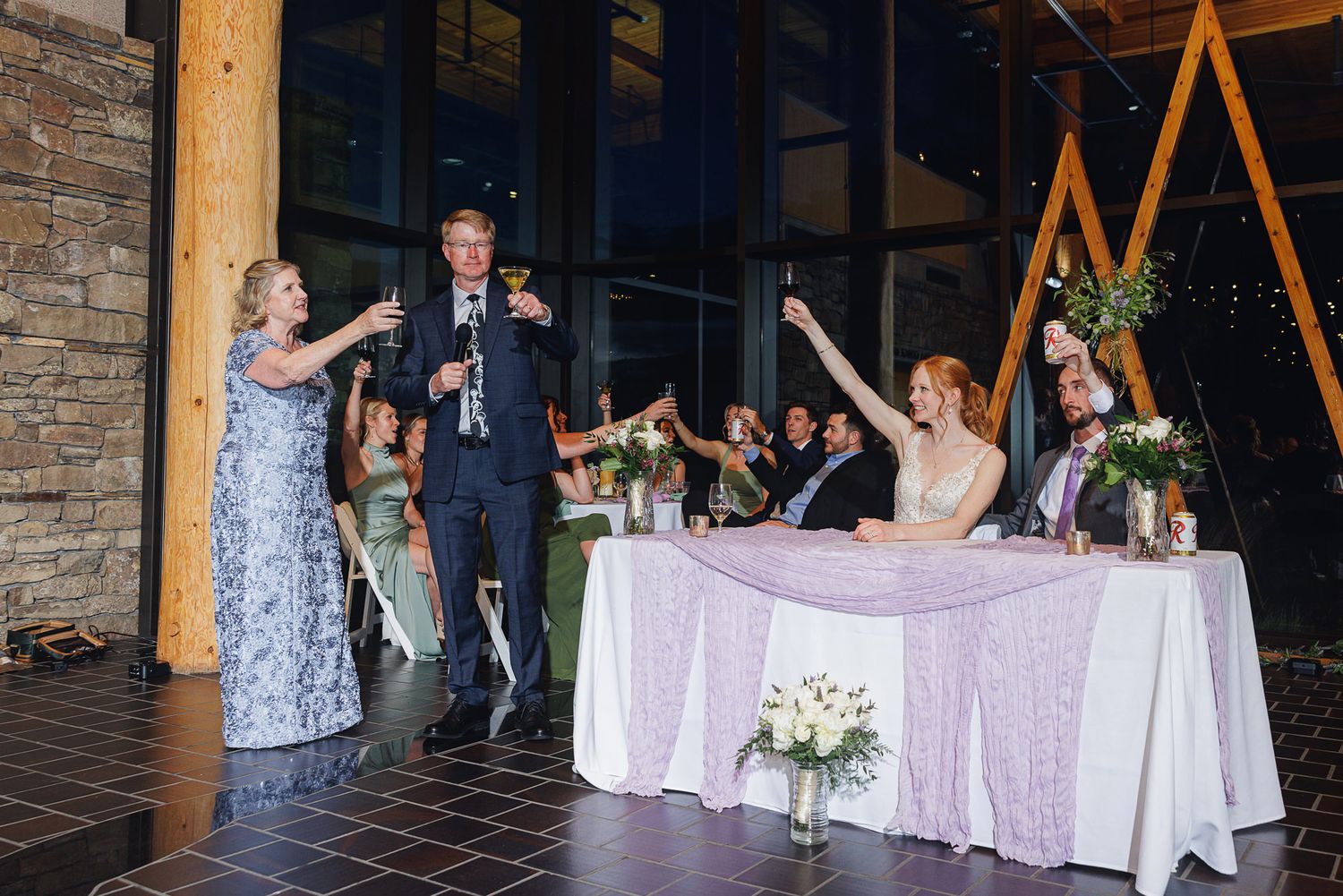 Wedding guests raise glasses in a toast to the bride and groom at an elegant reception venue.