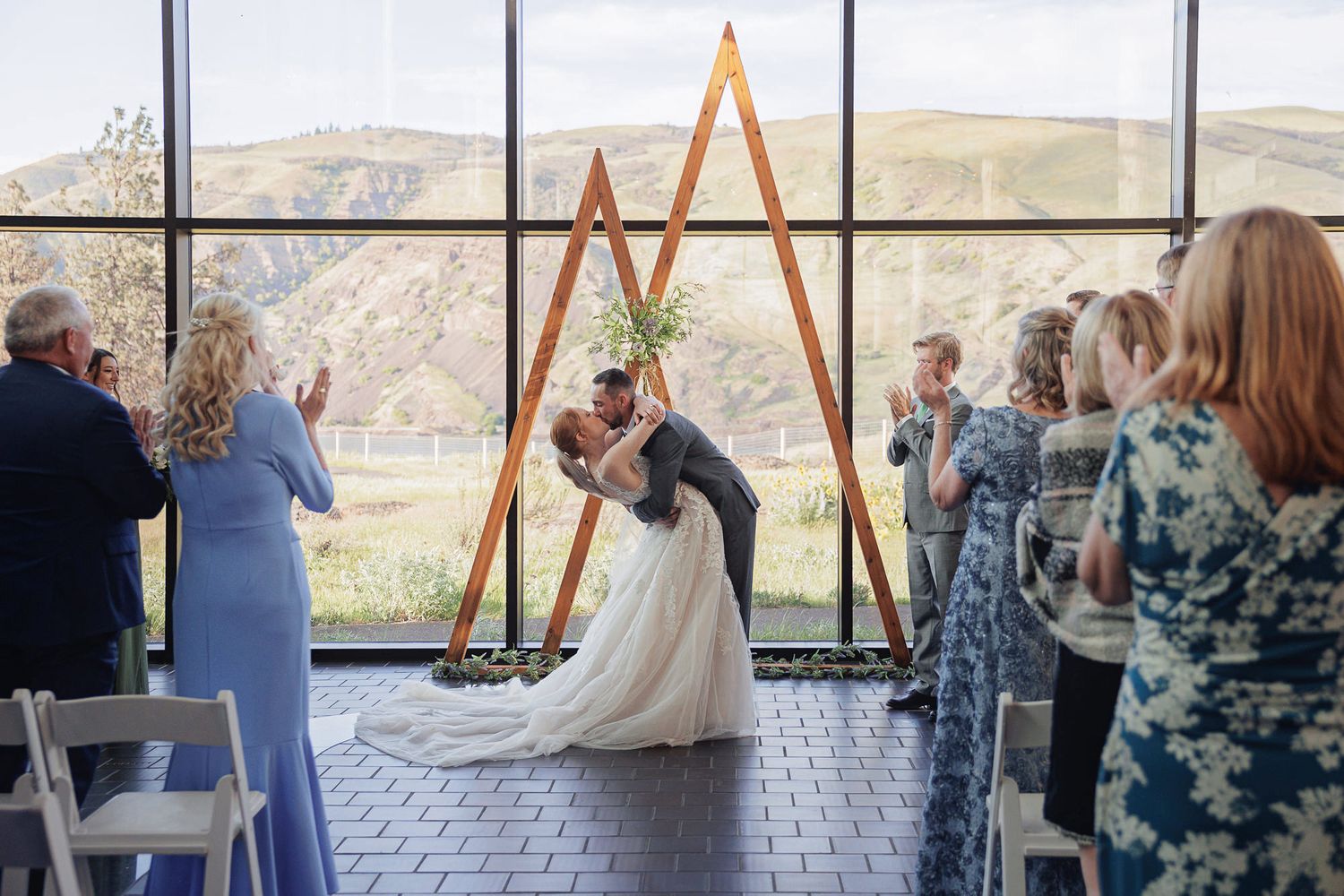 Bride and groom share first kiss at wedding ceremony with triangular wooden arch and mountain views.