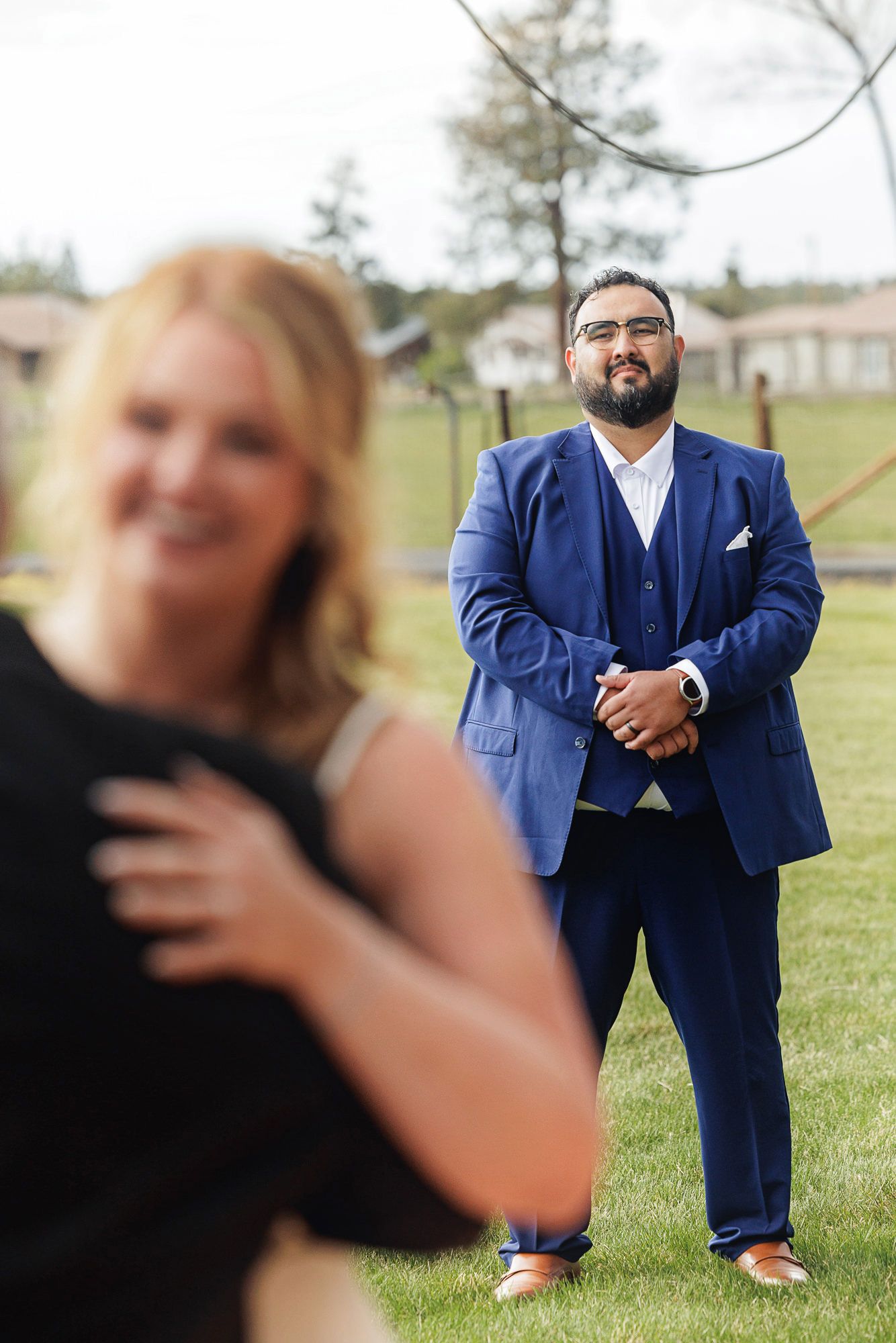 A man in a blue suit stands on green grass, gazing warmly at a blurred woman in the foreground at an outdoor event.
