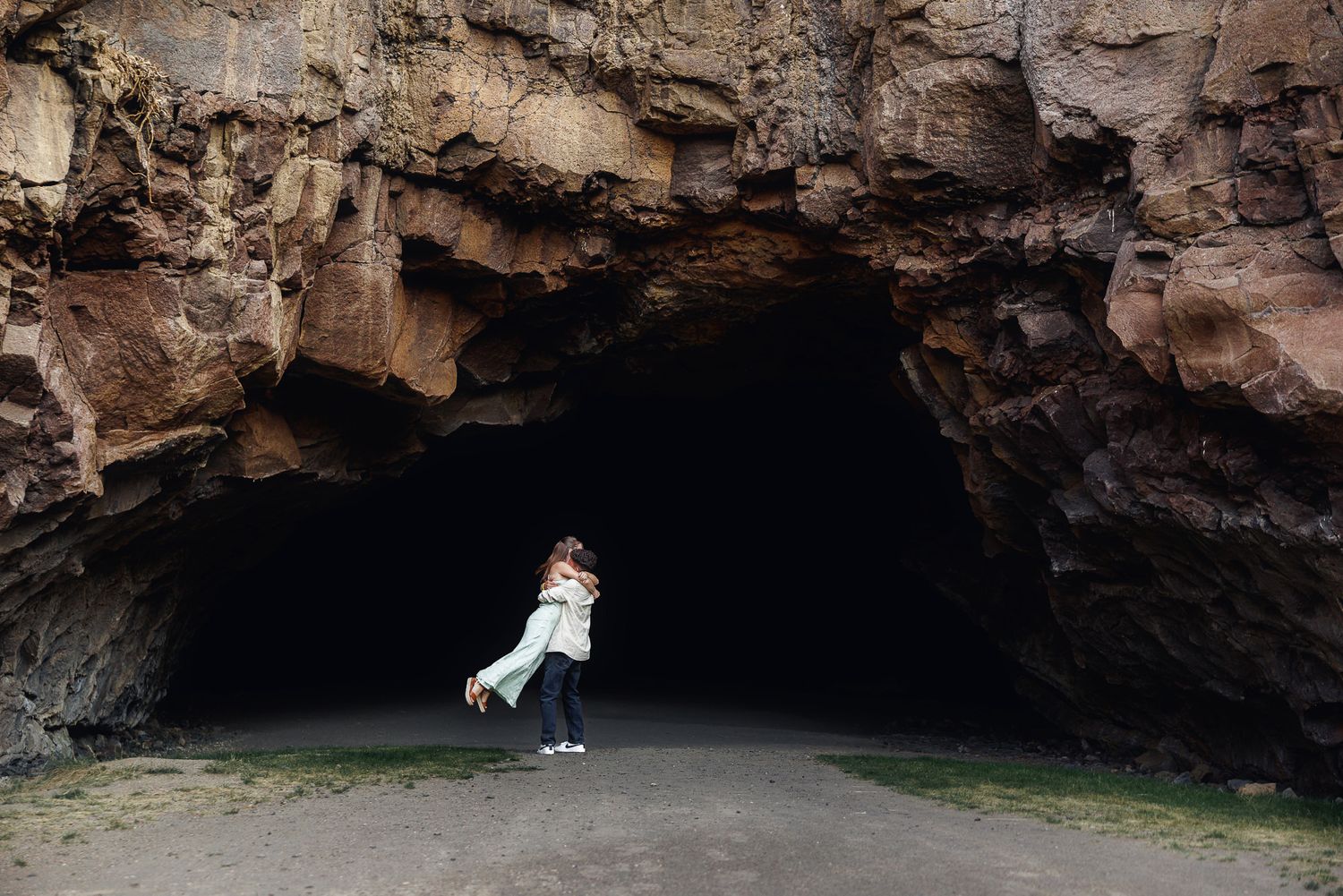 Couple embracing at the entrance of a dark sea cave with rocky walls on a coastal beach.