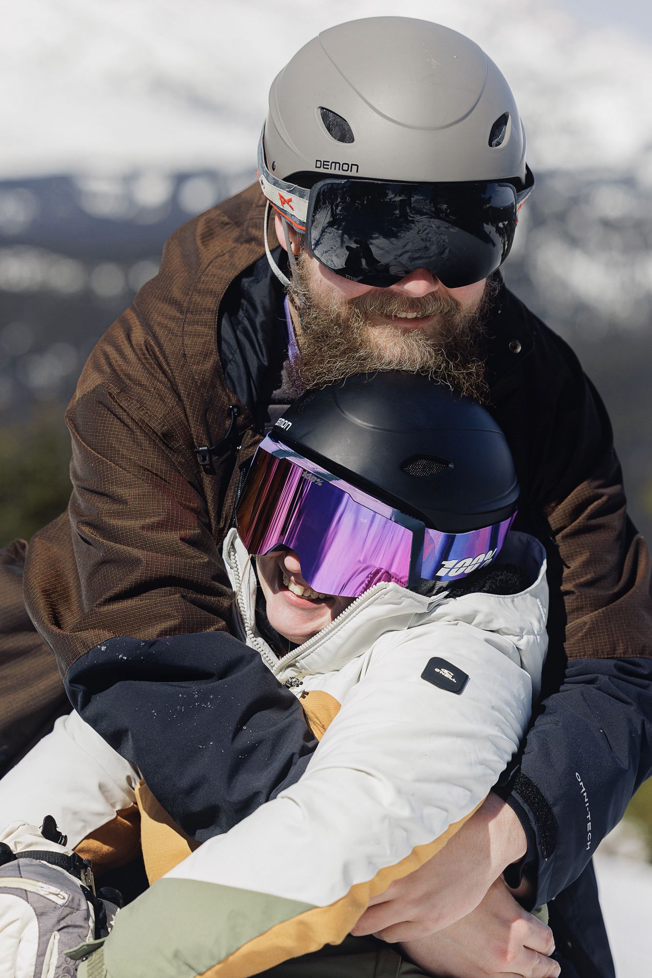 A bearded man wearing a ski helmet and goggles sits on a snowmobile in a snowy mountain landscape.