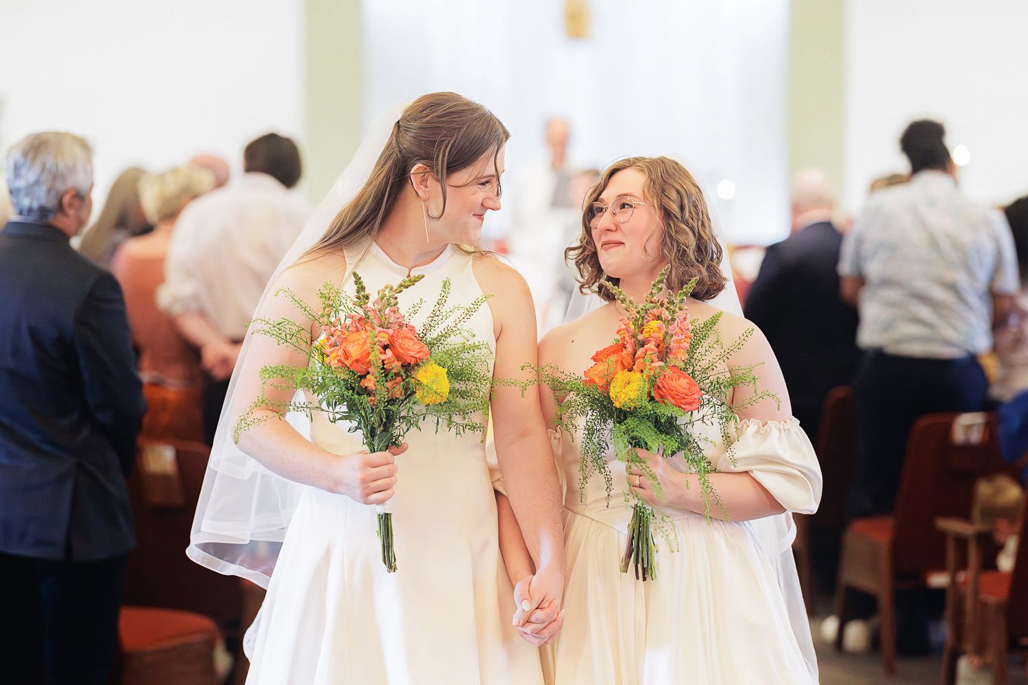 Two people in white dresses hold vibrant orange bouquets while sharing a joyful moment during a church ceremony.