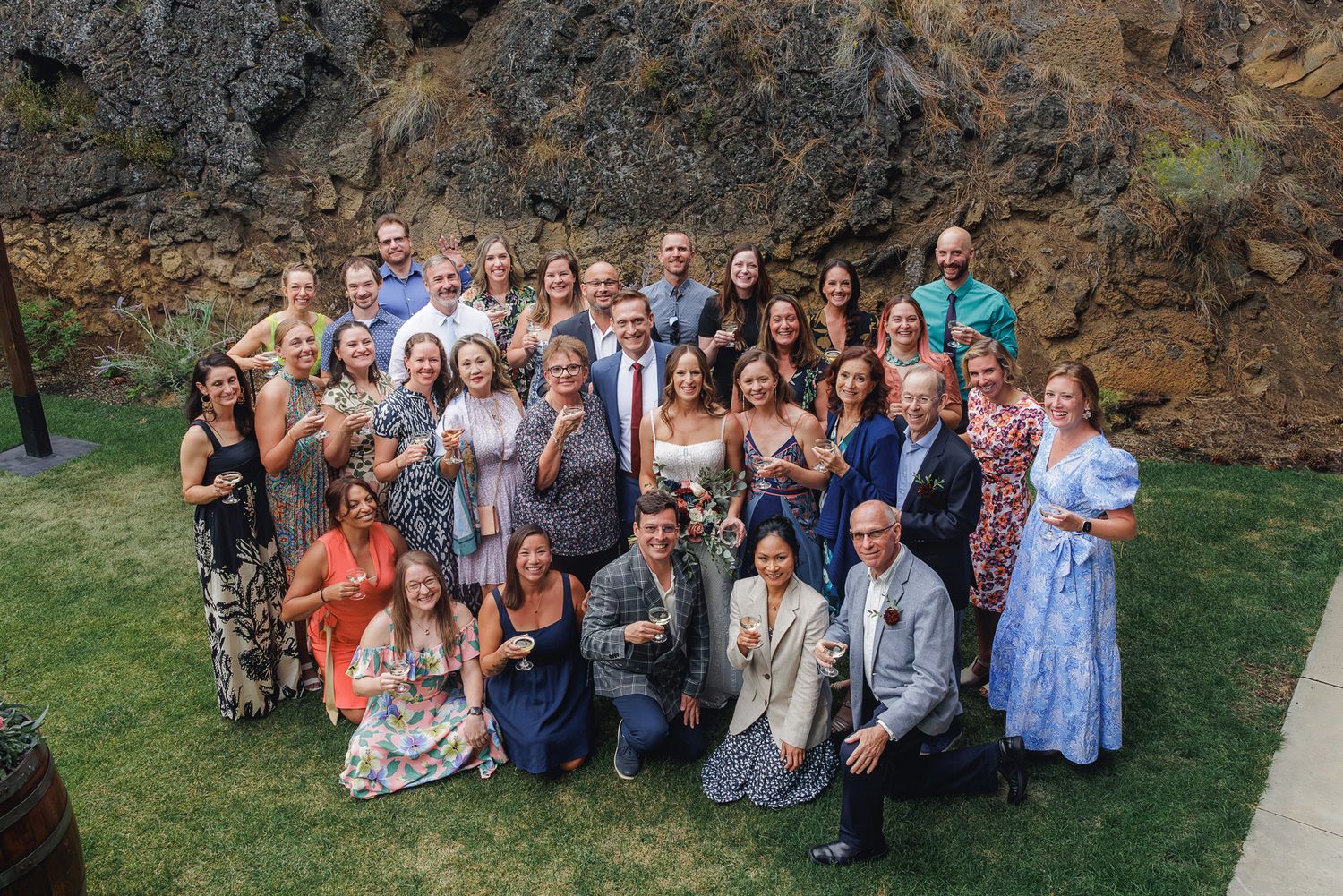 Large group of wedding guests gathered outdoors for panoramic photos against a natural backdrop.