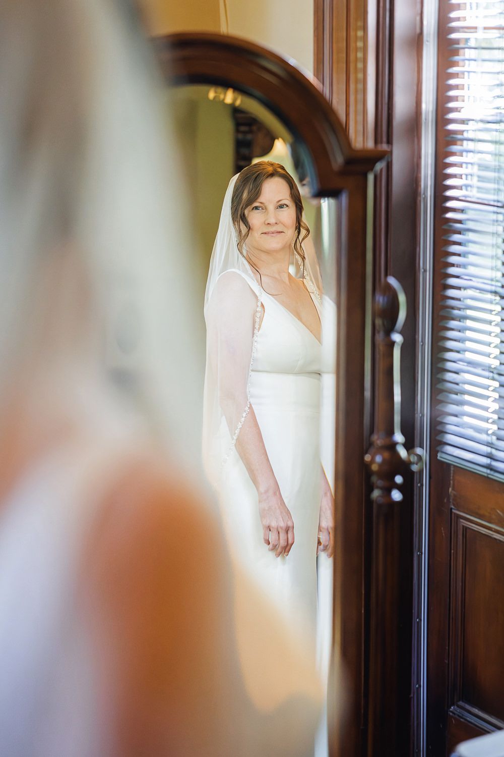 Person in a flowing white gown stands elegantly by a wooden doorway with window blinds visible in the background.