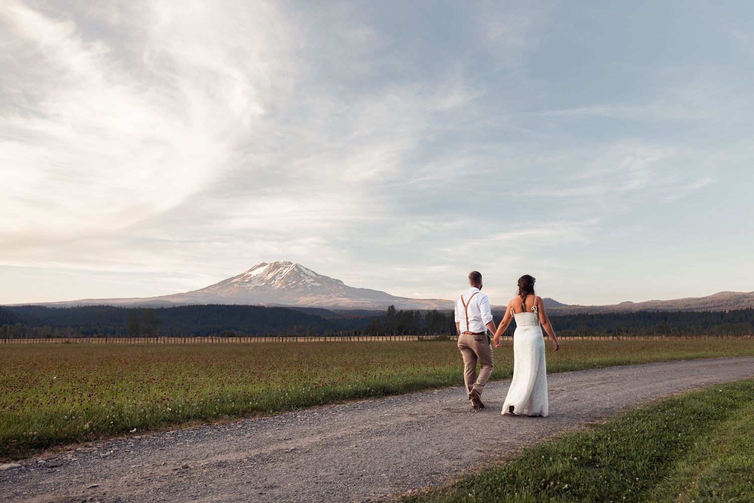 A couple in white wedding attire walks along a country road with snow-capped Mount Hood in the distance.