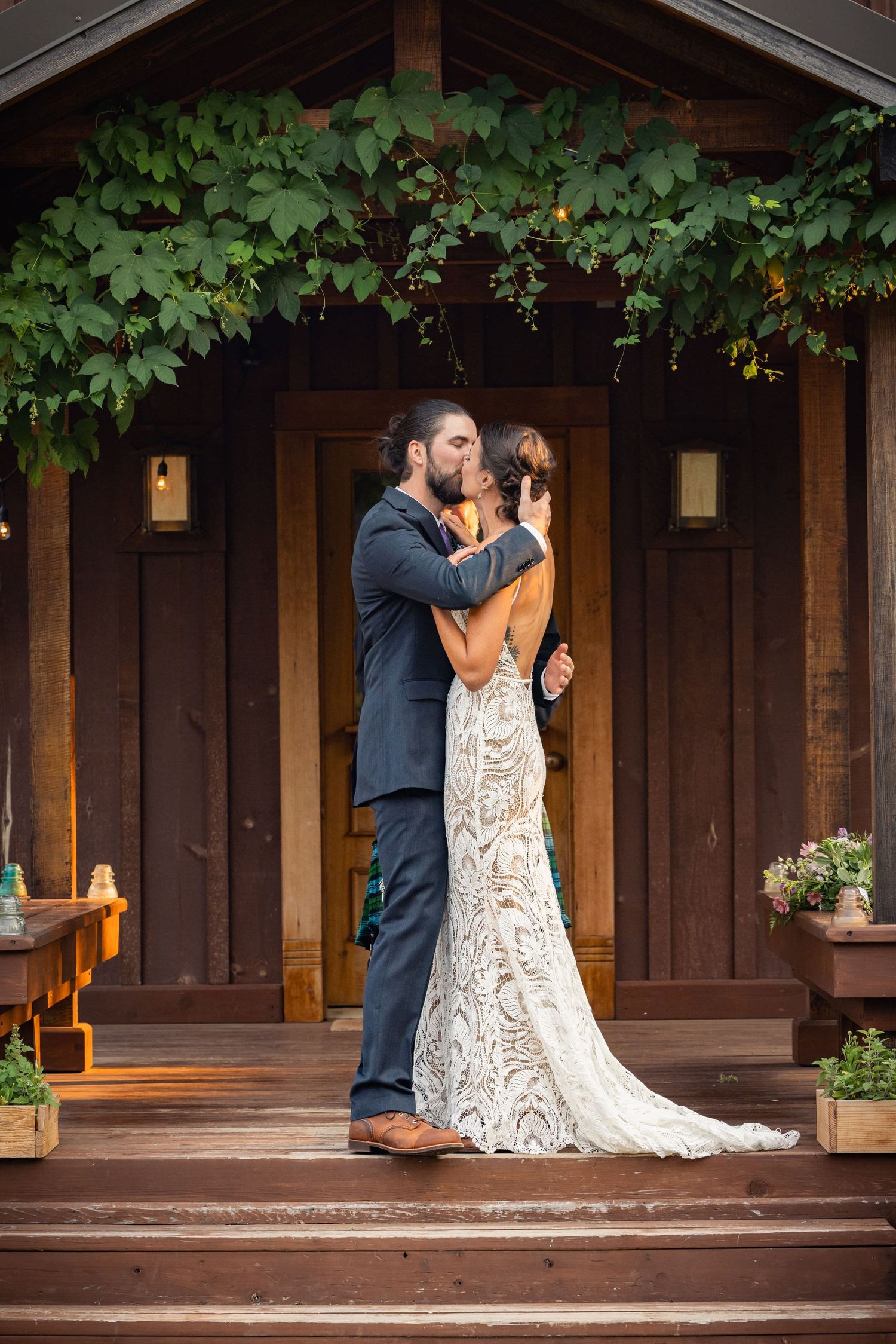 A couple embraces on wooden steps outside a rustic cabin with greenery hanging above the entrance.
