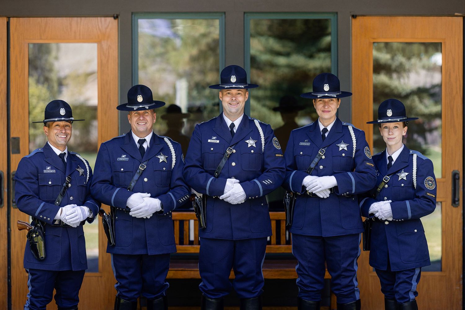 Five police officers in blue uniforms and white gloves stand at attention in front of glass doors.
