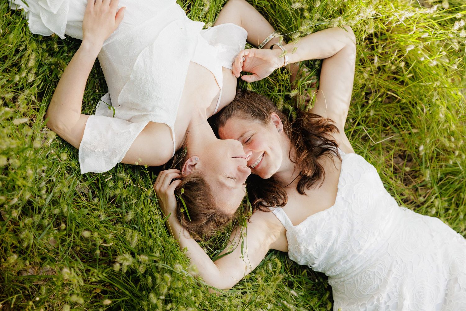 Two people in white dresses lay in lush green grass creating a romantic outdoor portrait.