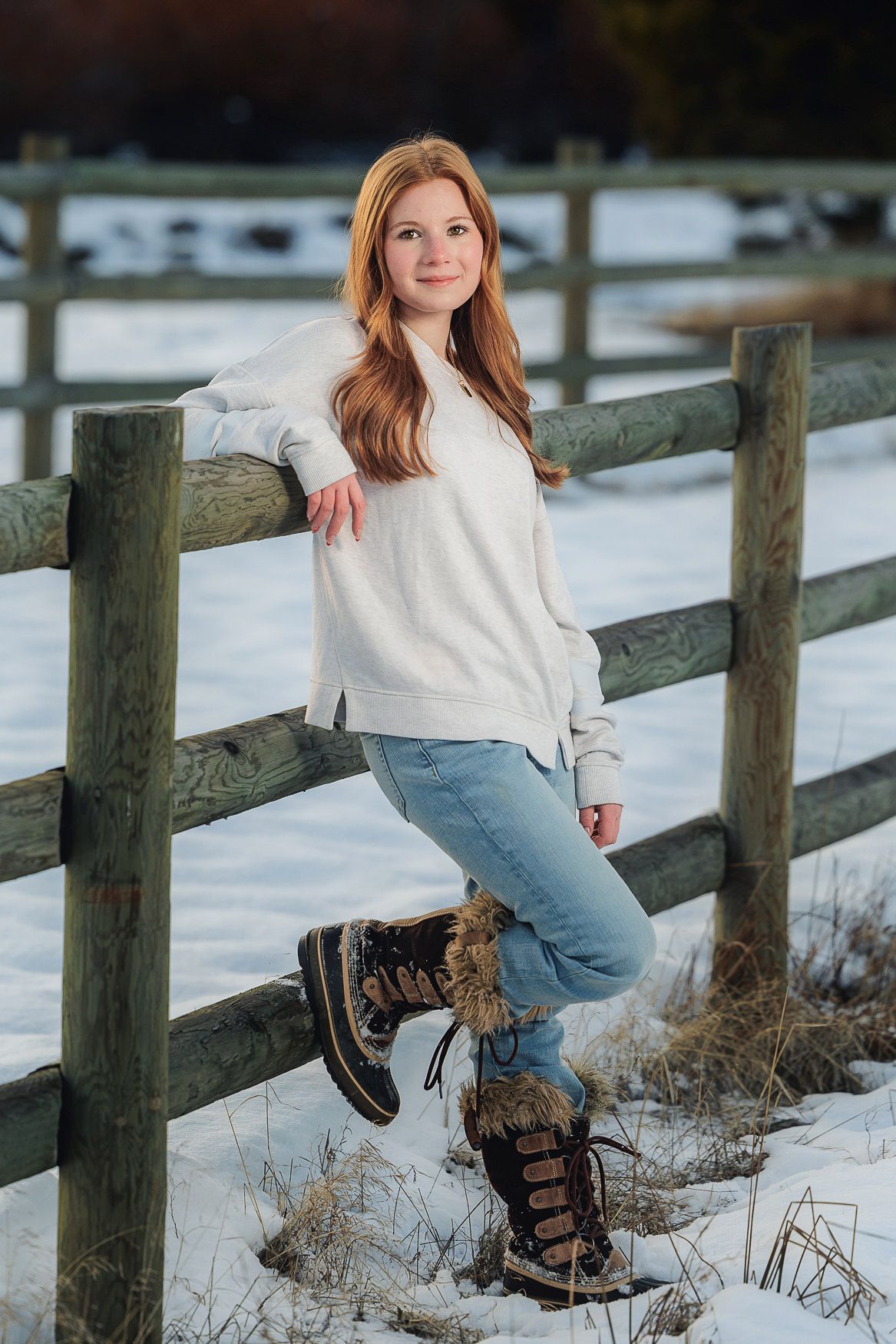 Winter portrait of someone in white sweater and snow boots leaning against wooden fence in snow.