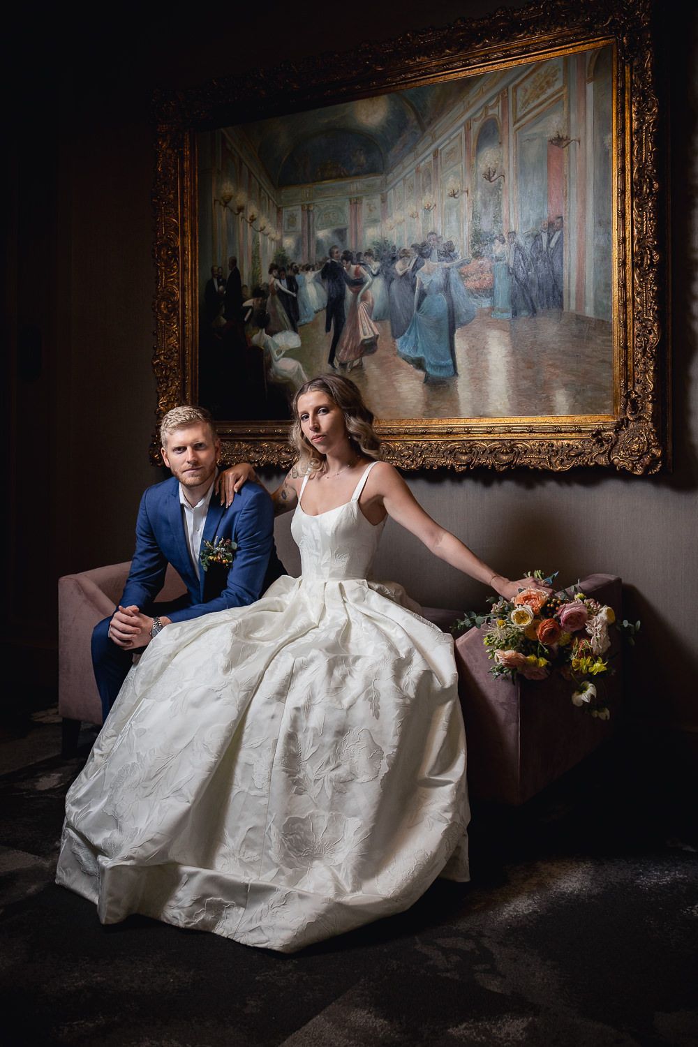 A couple dressed in formal wedding attire sits on a couch beneath an ornate gold-framed painting depicting a ballroom scene.