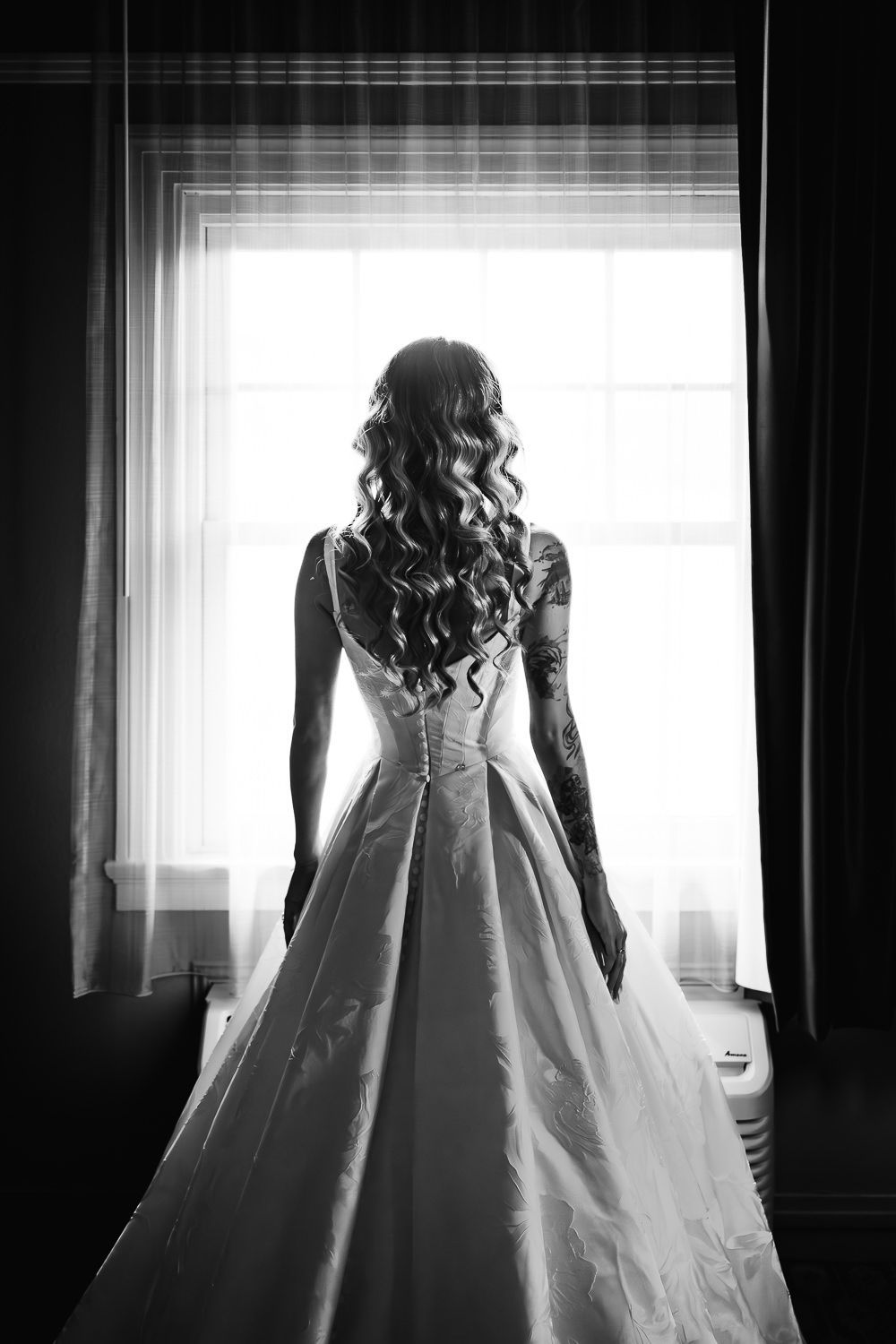 Black and white silhouette of a bride in a wedding gown with long curled hair standing by a bright window.