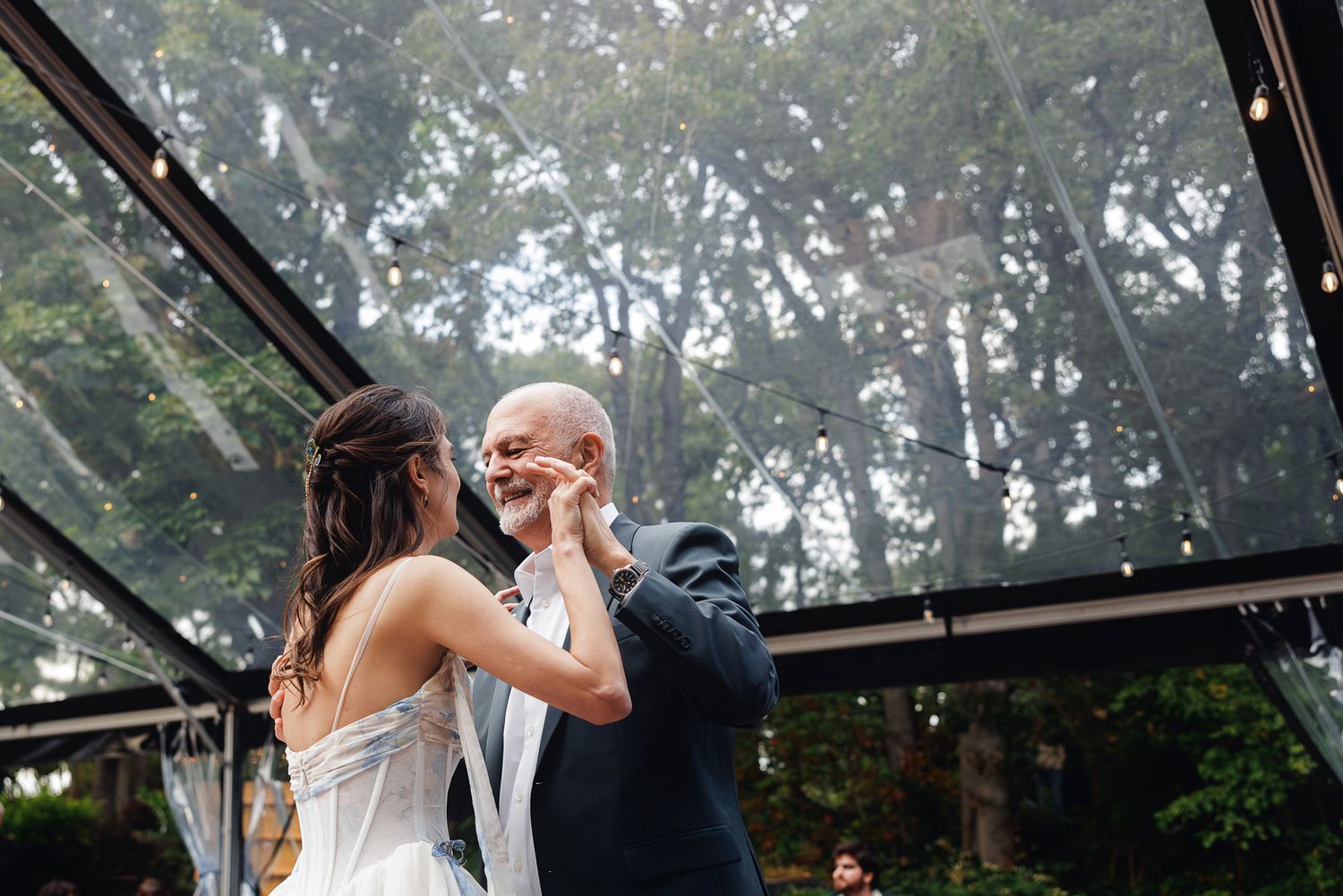 Bride and groom share an emotional first dance under a glass ceiling tent surrounded by lush trees at an outdoor wedding.