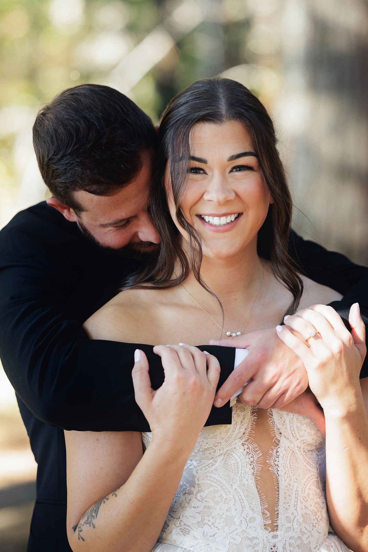 A smiling bride in a lace gown embraced from behind by her groom in black, showcasing her engagement ring.
