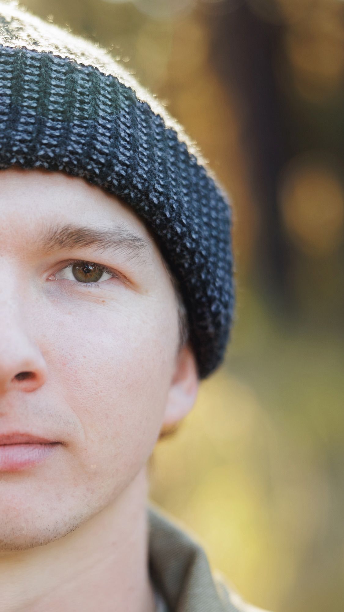 Close-up half portrait of a young man in a dark knit beanie and olive jacket against a softly blurred autumn forest background.