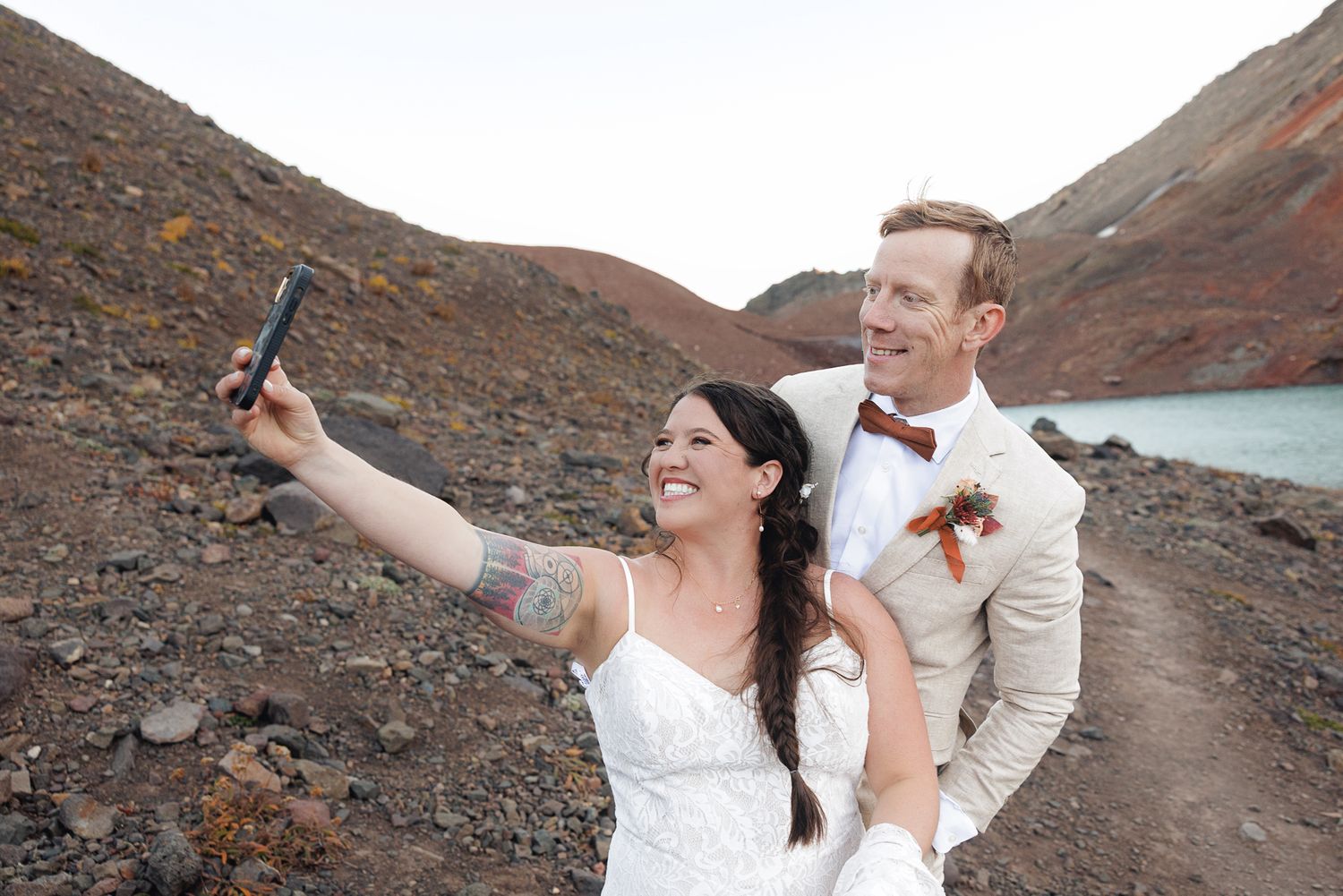 Smiling bride in a white dress and groom in a beige suit take a selfie on a rocky volcanic trail beside a turquoise lake.