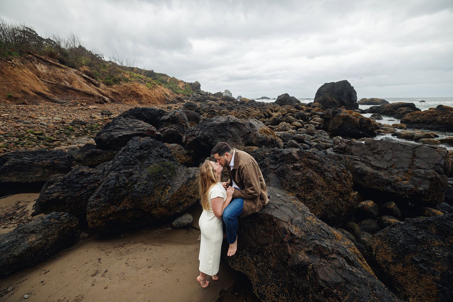 Romantic couple sharing a kiss on coastal rocks at a cloudy beach.