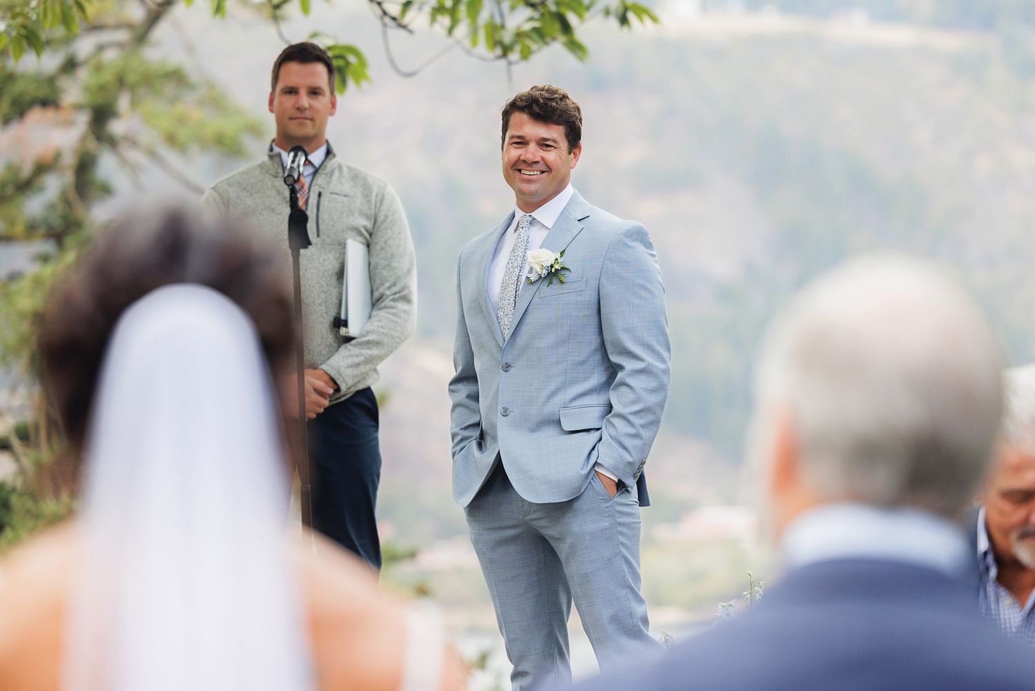 Wedding ceremony guests seated outdoors on white chairs with mountain views and greenery in the background.