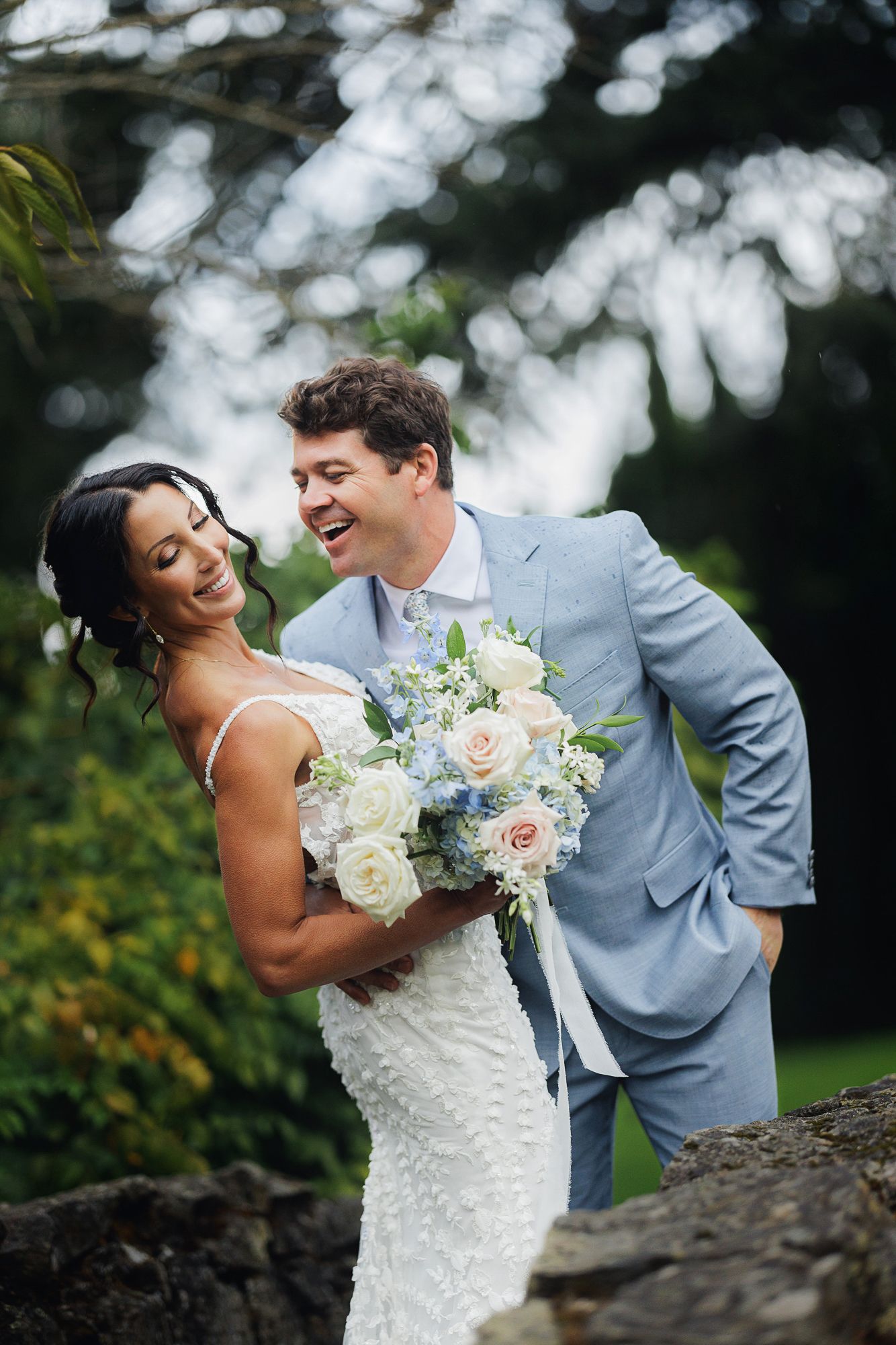 A joyful wedding couple shares a candid moment outdoors with white flowers and bokeh lighting in the background.