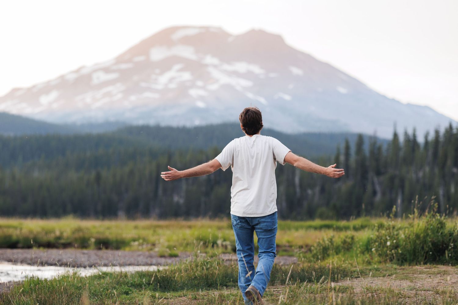 Someone stands with outstretched arms in a meadow overlooking a snow-capped mountain peak and evergreen forest.