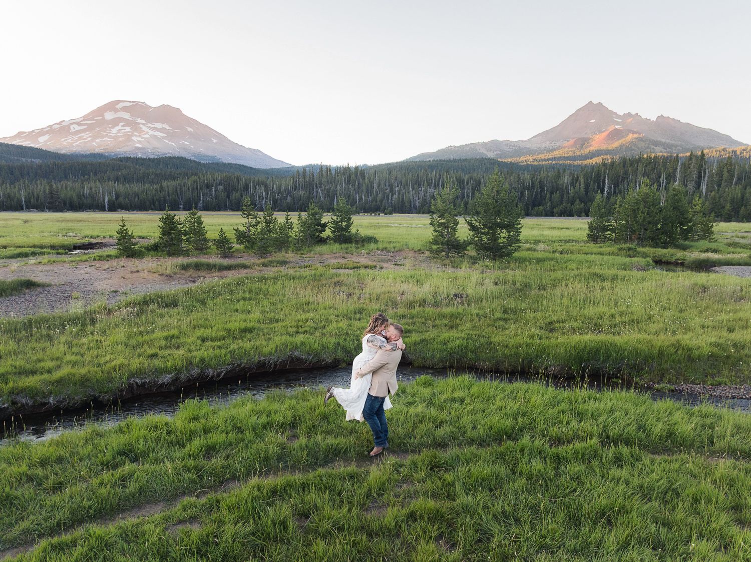 Person dancing in a meadow with winding creek and mountain peaks in the background under cloudy skies.