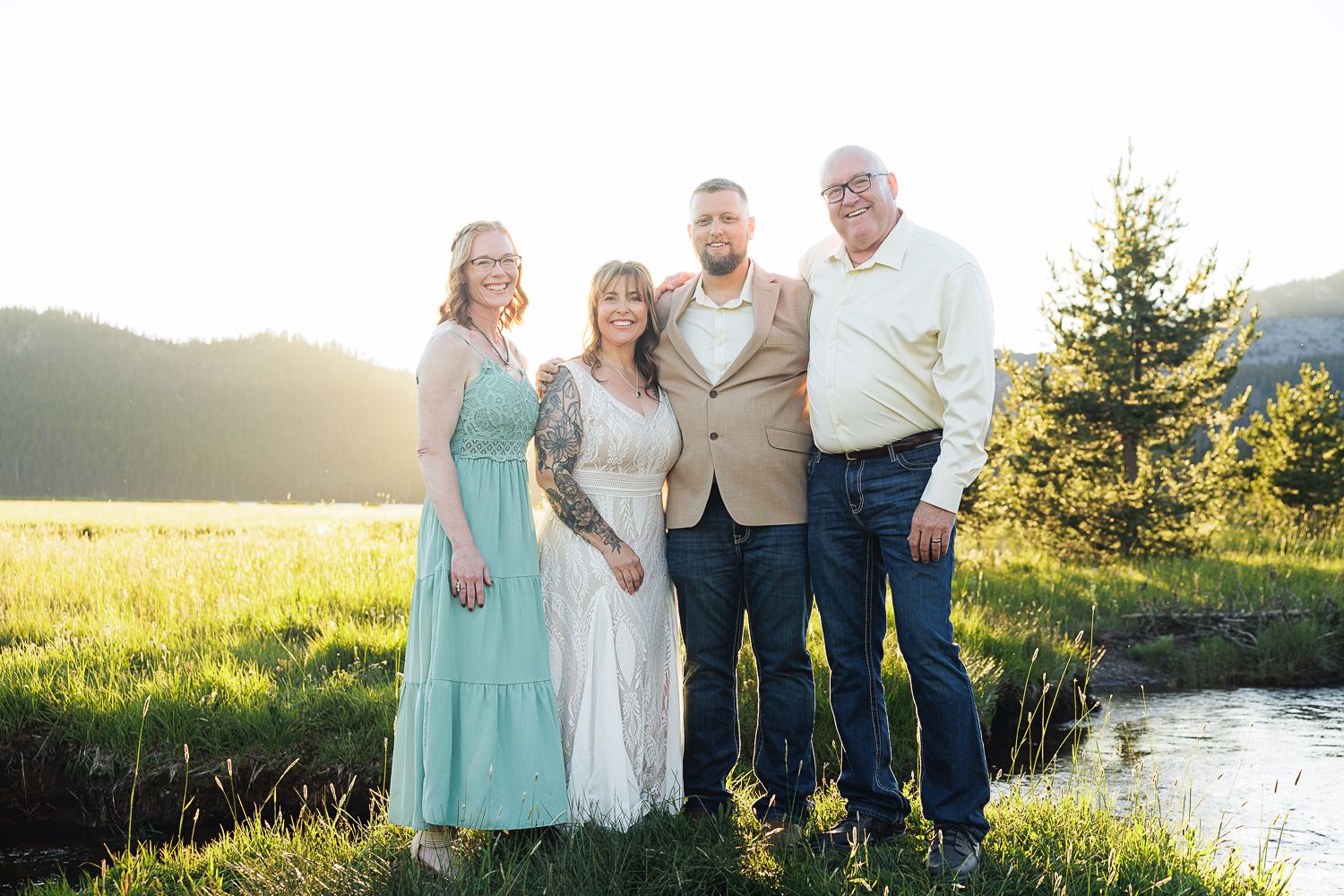 A group gathers together on a sun-drenched meadow near a stream with mountains and evergreen trees in the background.