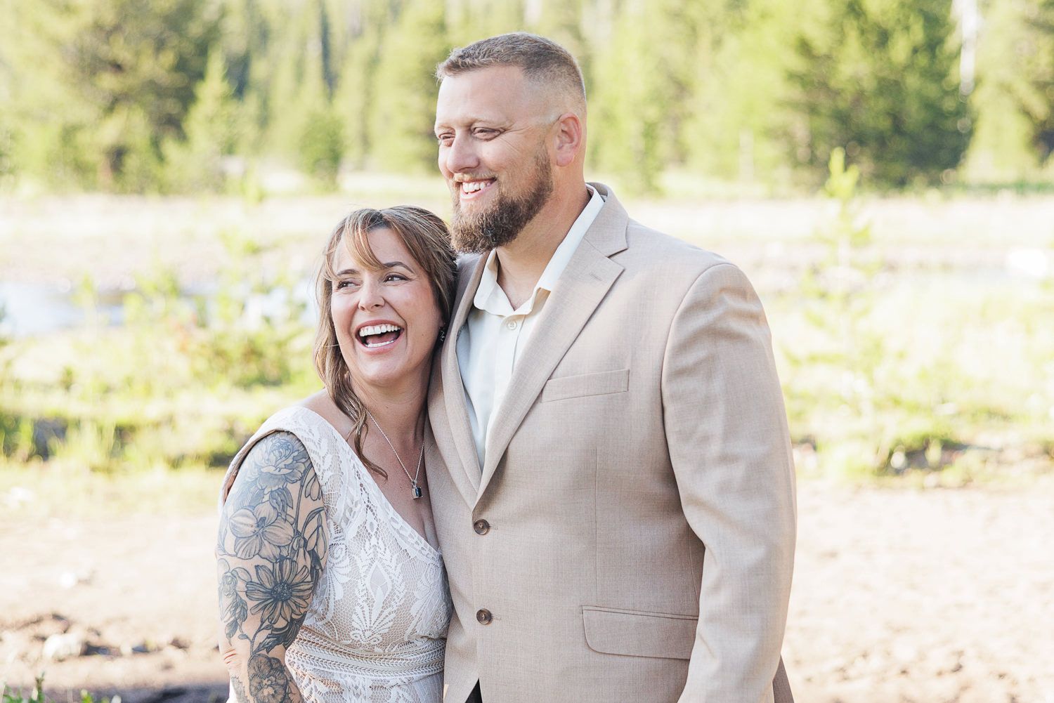 A happy couple poses outdoors in wedding attire, with the bride in a lace dress and groom in a beige suit against a natural backdrop.