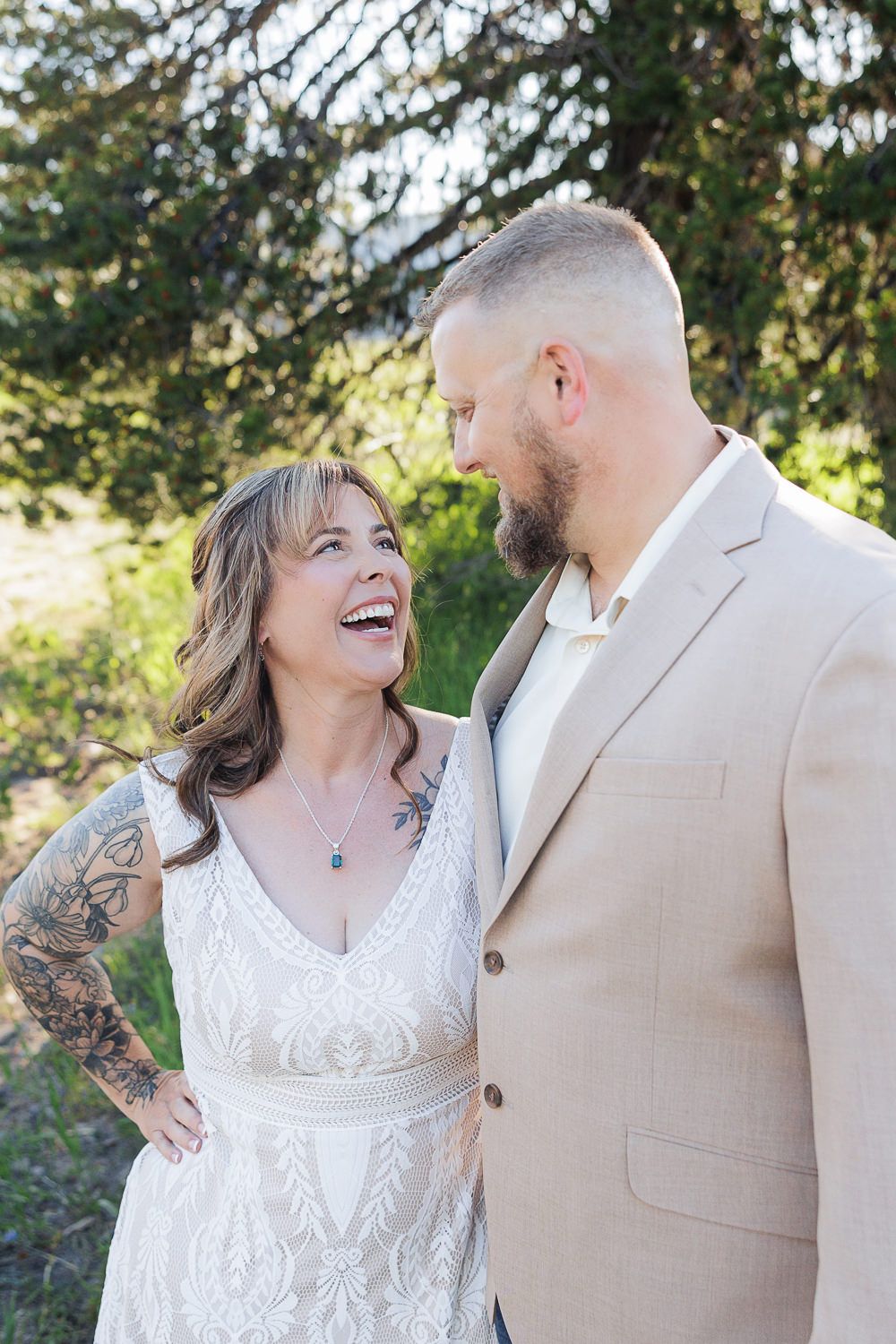 A couple in wedding attire share joyful laughter outdoors, with the bride in white lace and groom in tan suit.