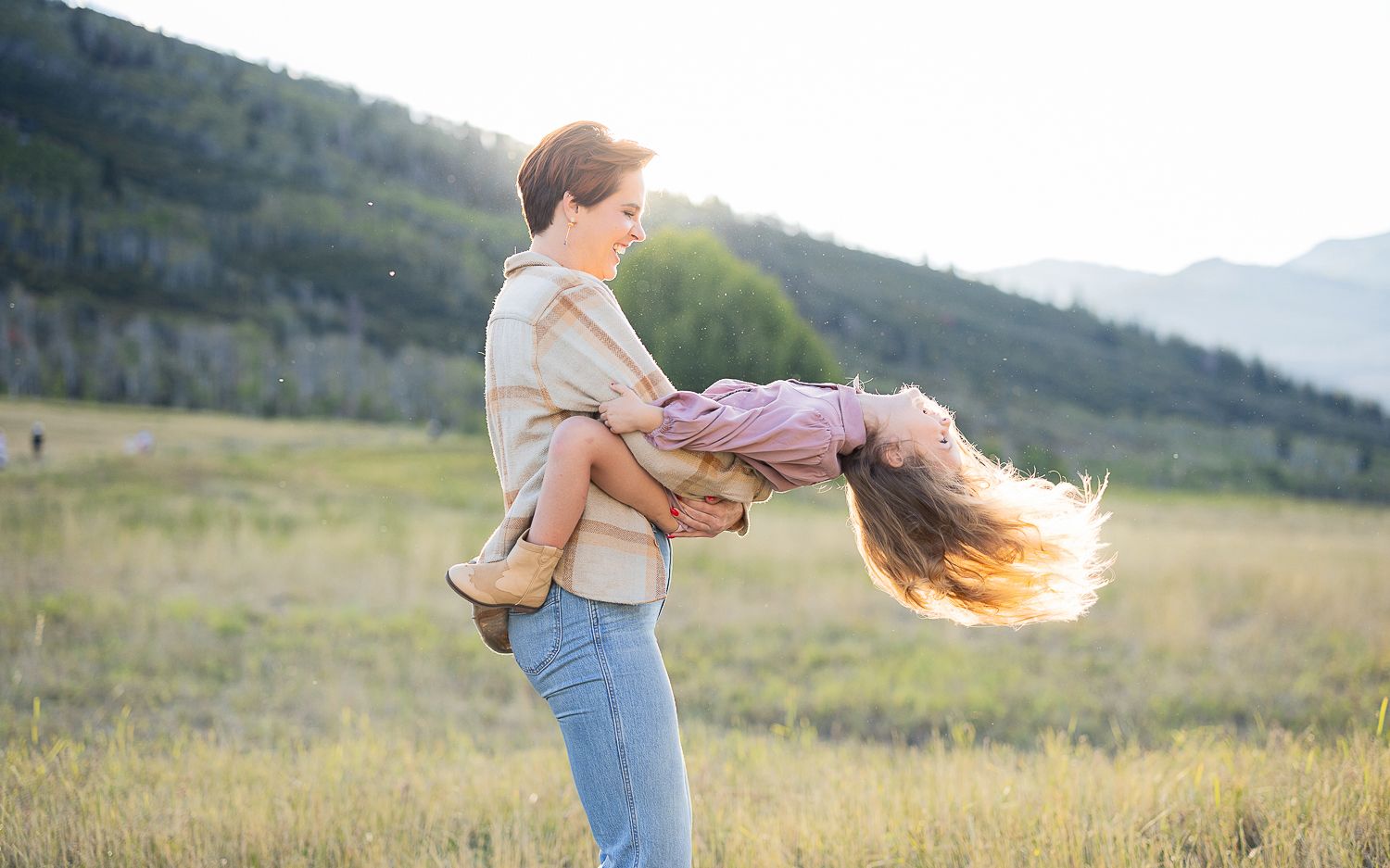 A playful couple dance together in a golden meadow at sunset with mountains in the background.