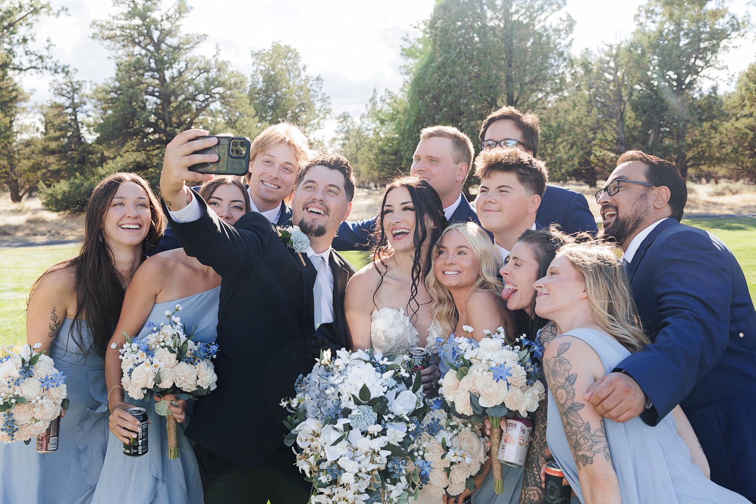 Wedding party in light blue dresses and dark suits pose together outdoors while laughing and smiling.