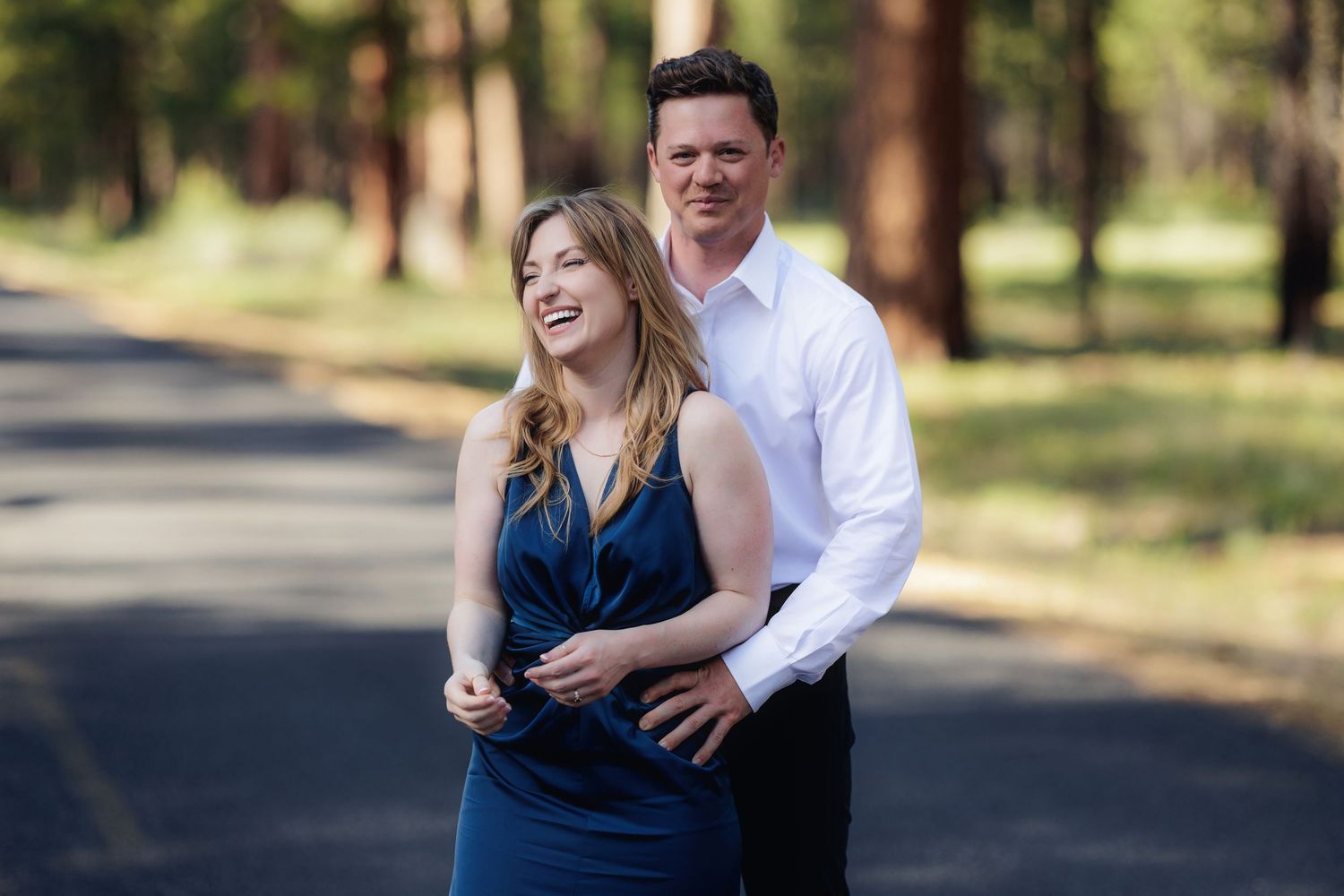 A romantic couple laughs and embraces while walking together on a sun-dappled pathway through a park.
