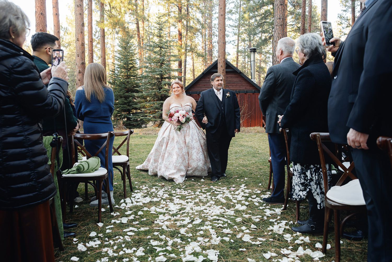 Wedding guests gather for an intimate outdoor ceremony surrounded by tall pine trees and scattered white flower petals.