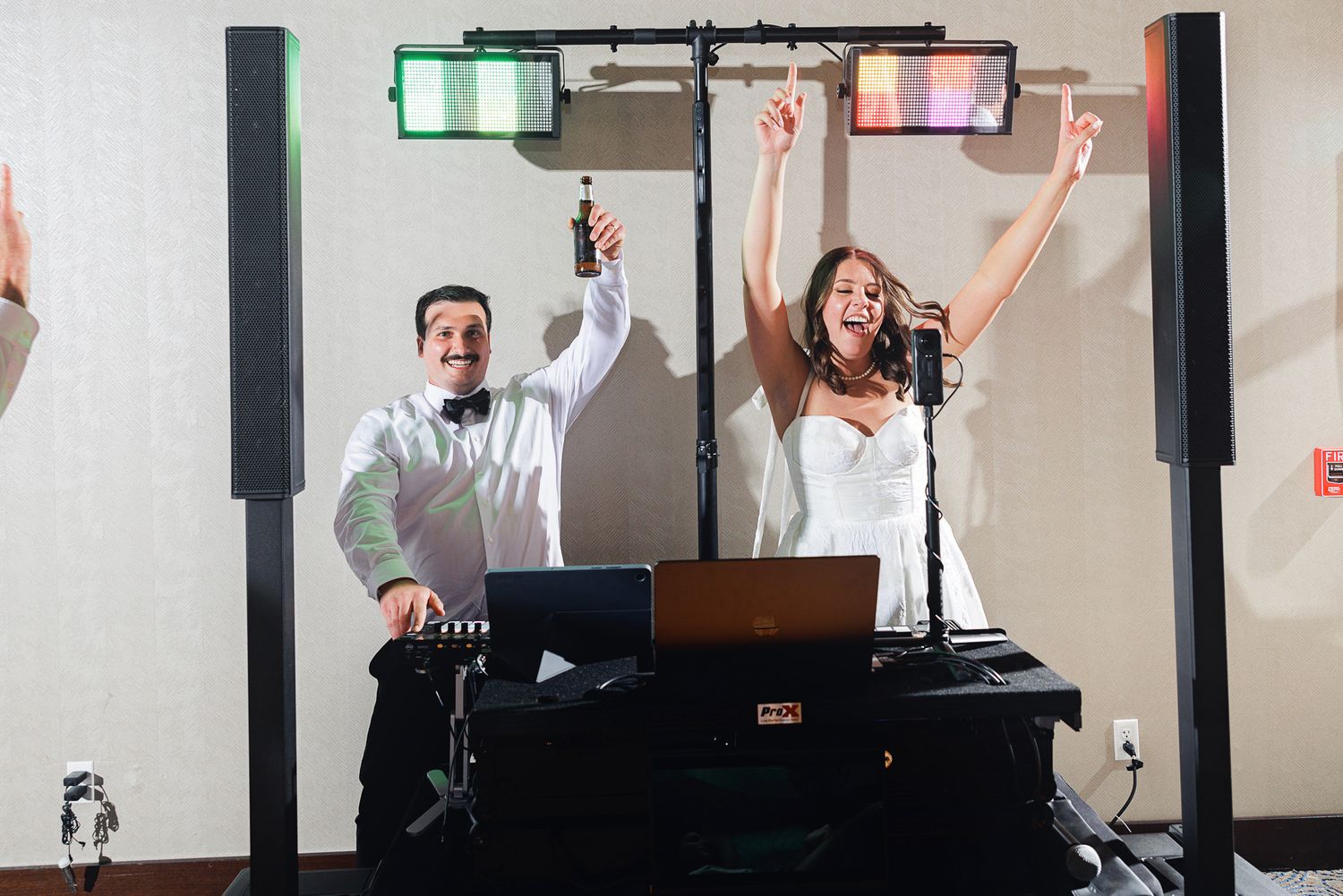 Bride and groom celebrate behind DJ booth with colorful lights at lively wedding reception.